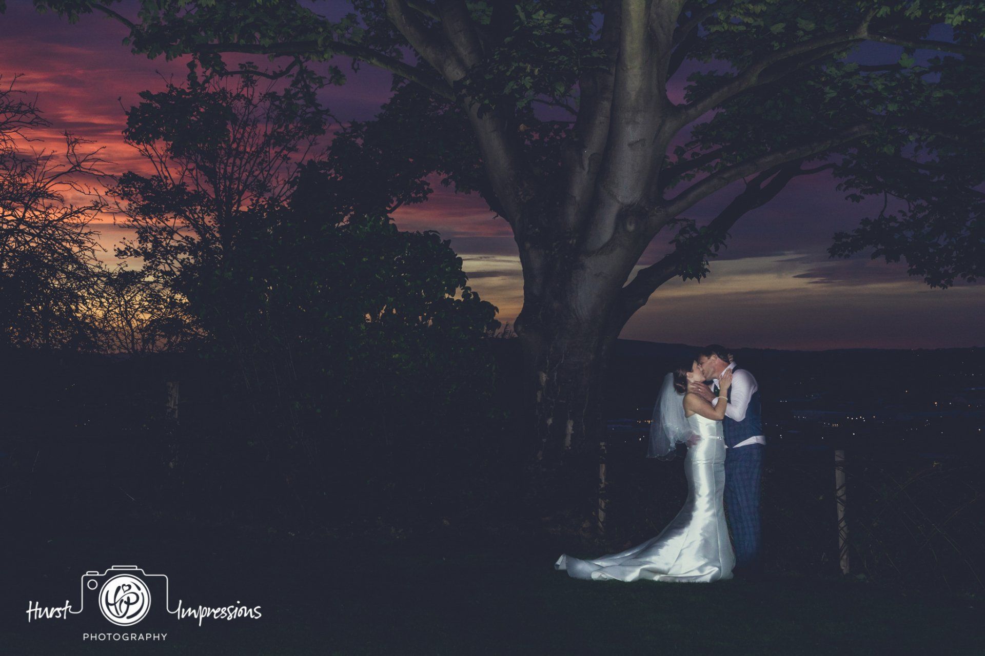 Bride and groom kissing under a tree at sunset. Vibrant orange and purple sky.