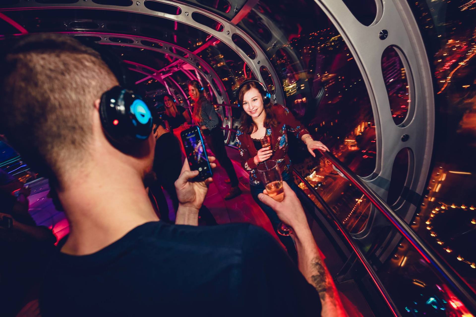 People at a silent disco on a high walkway with city views. Headphones on, some holding drinks.