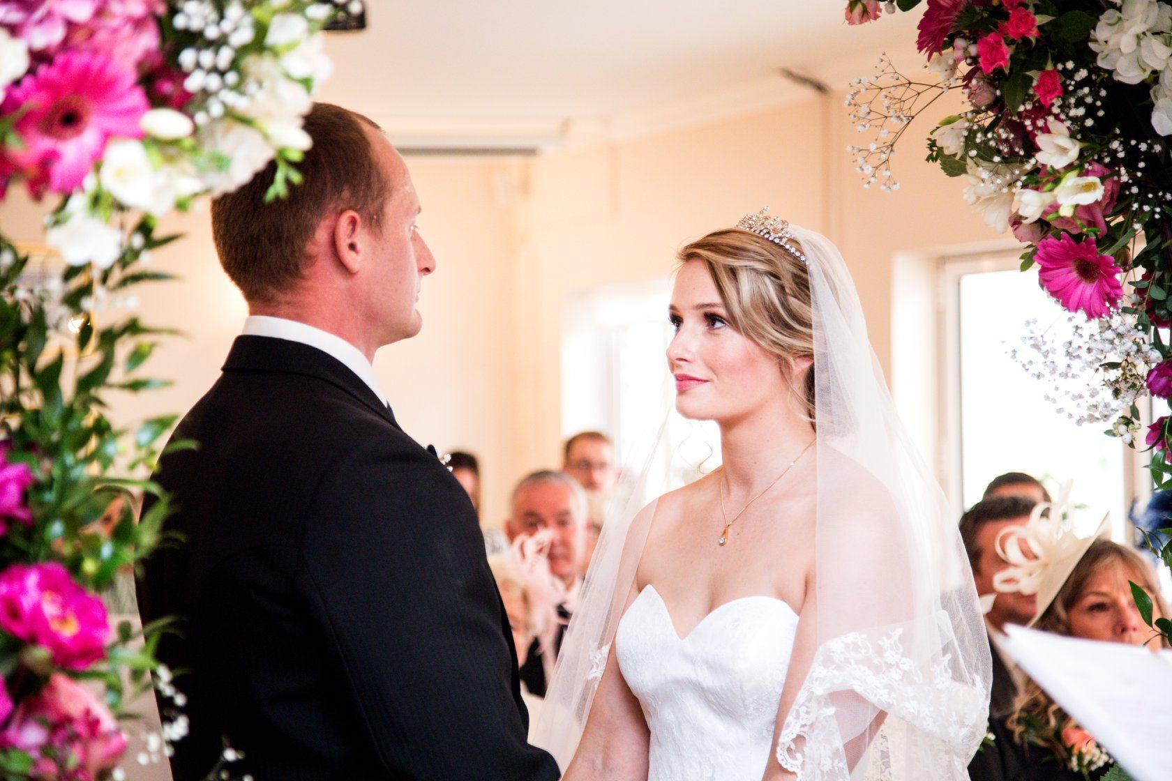 Bride and groom at altar under floral arch; she gazes at him, he faces her. Guests watch.