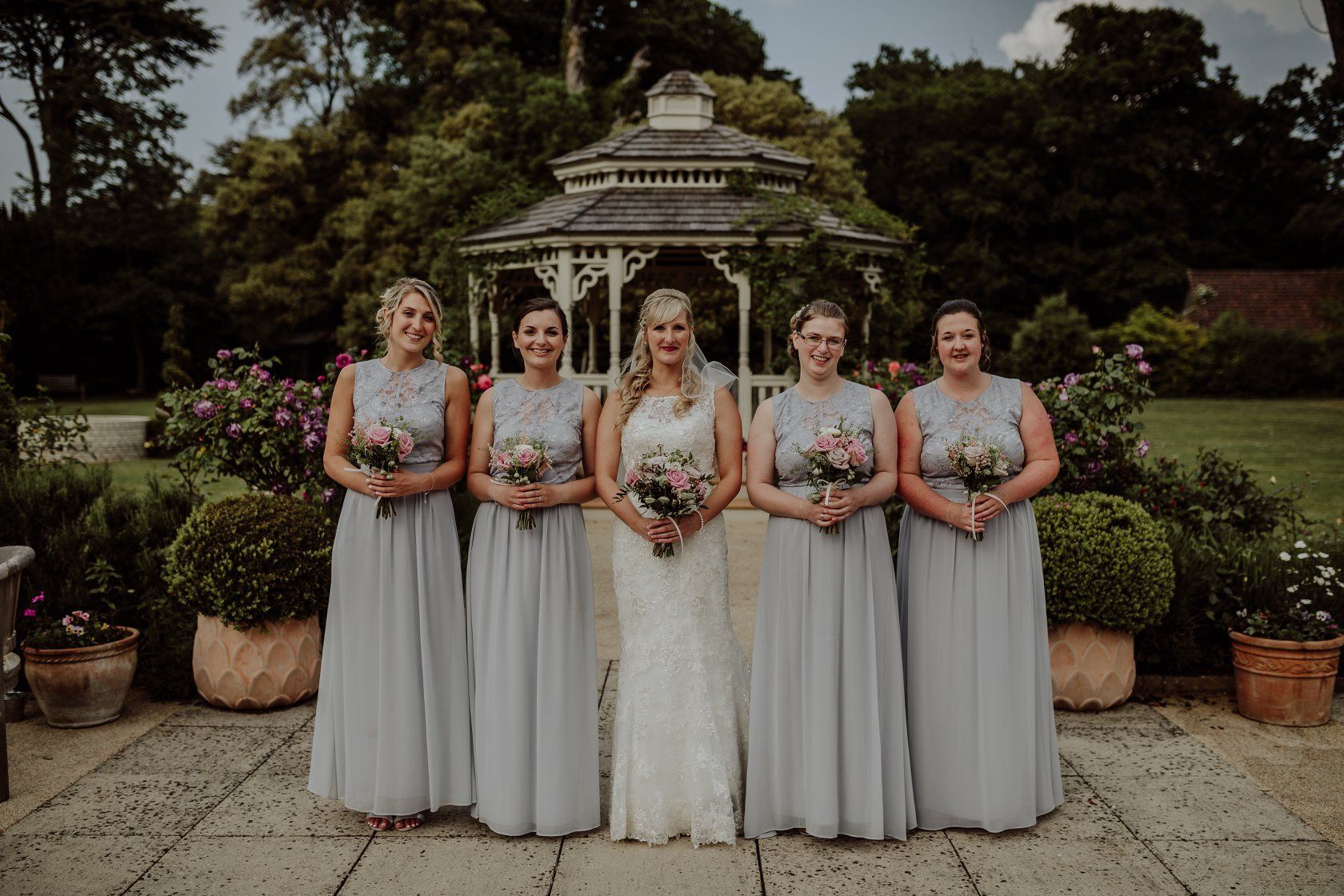 Bride with bridesmaids, wearing grey dresses, pose in front of a gazebo and garden.