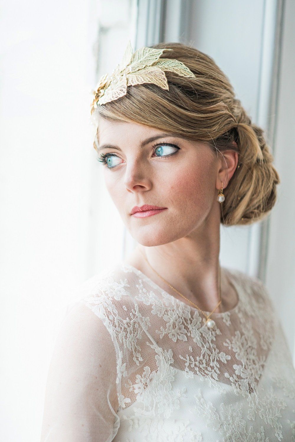 Woman in a white lace dress with a gold headpiece, looking out a window.