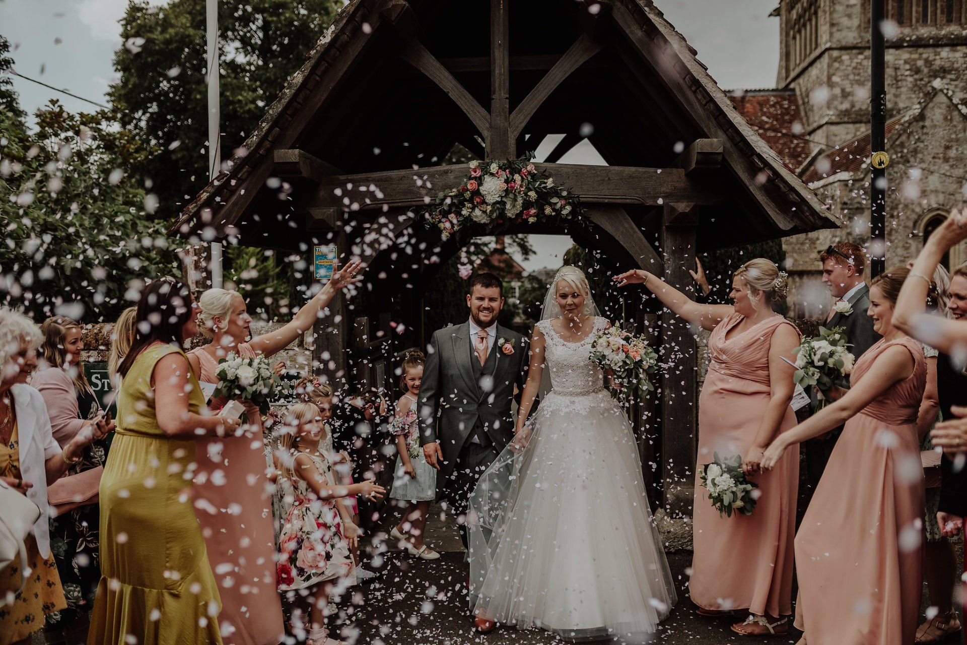 Newlyweds exit a church, showered with confetti by smiling wedding guests.