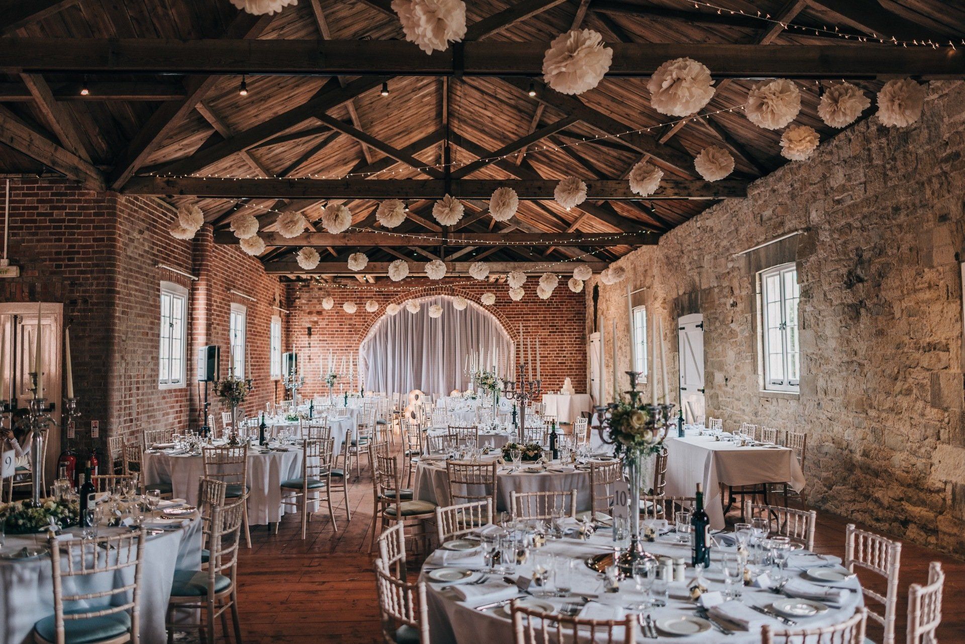 Wedding reception in a rustic hall with round tables, white tablecloths, and paper decorations hanging from the wooden ceiling.
