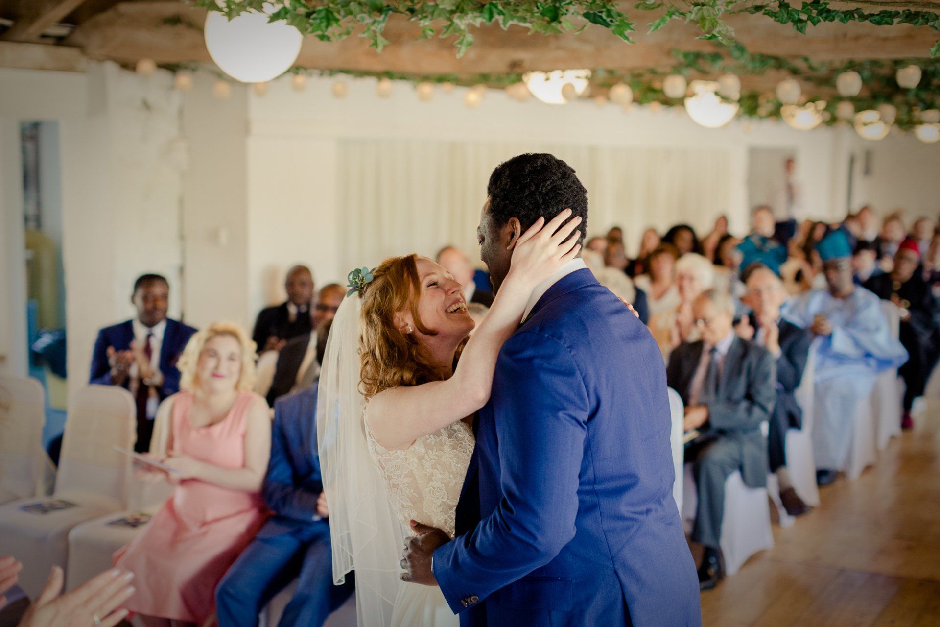 Newlyweds embrace, celebrating amidst wedding guests, indoor ceremony, white and blue attire.