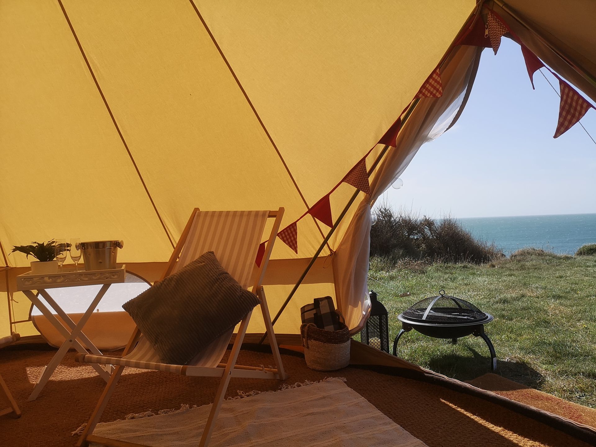 View from inside a canvas tent towards ocean. Chairs, a small table, and fire pit are visible.