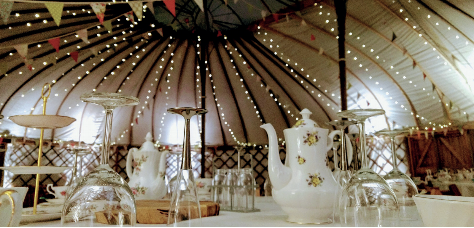 Inside a yurt, a table set with teacups, teapot, and glassware, strung with decorative lights and bunting.