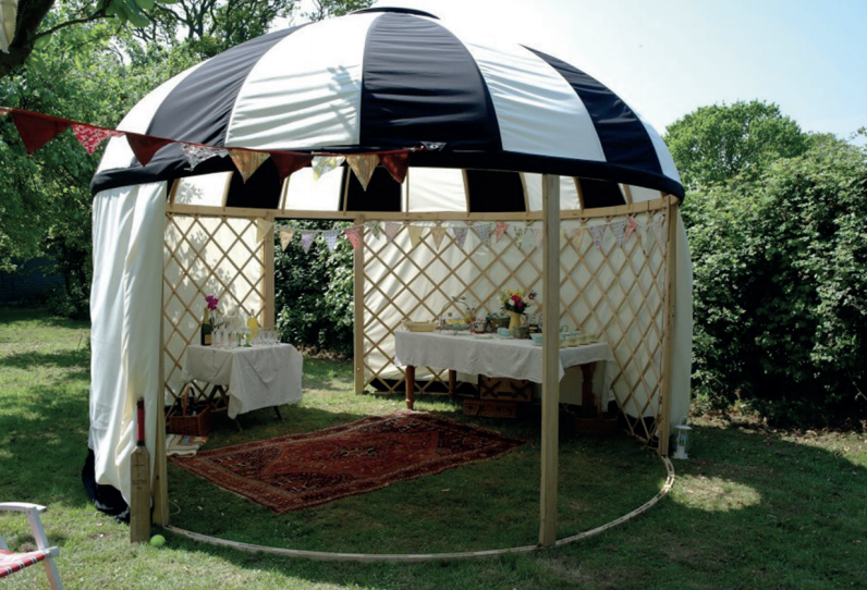 Circular, black-and-white striped tent with a wooden frame in a grassy yard, with tables set for a gathering.