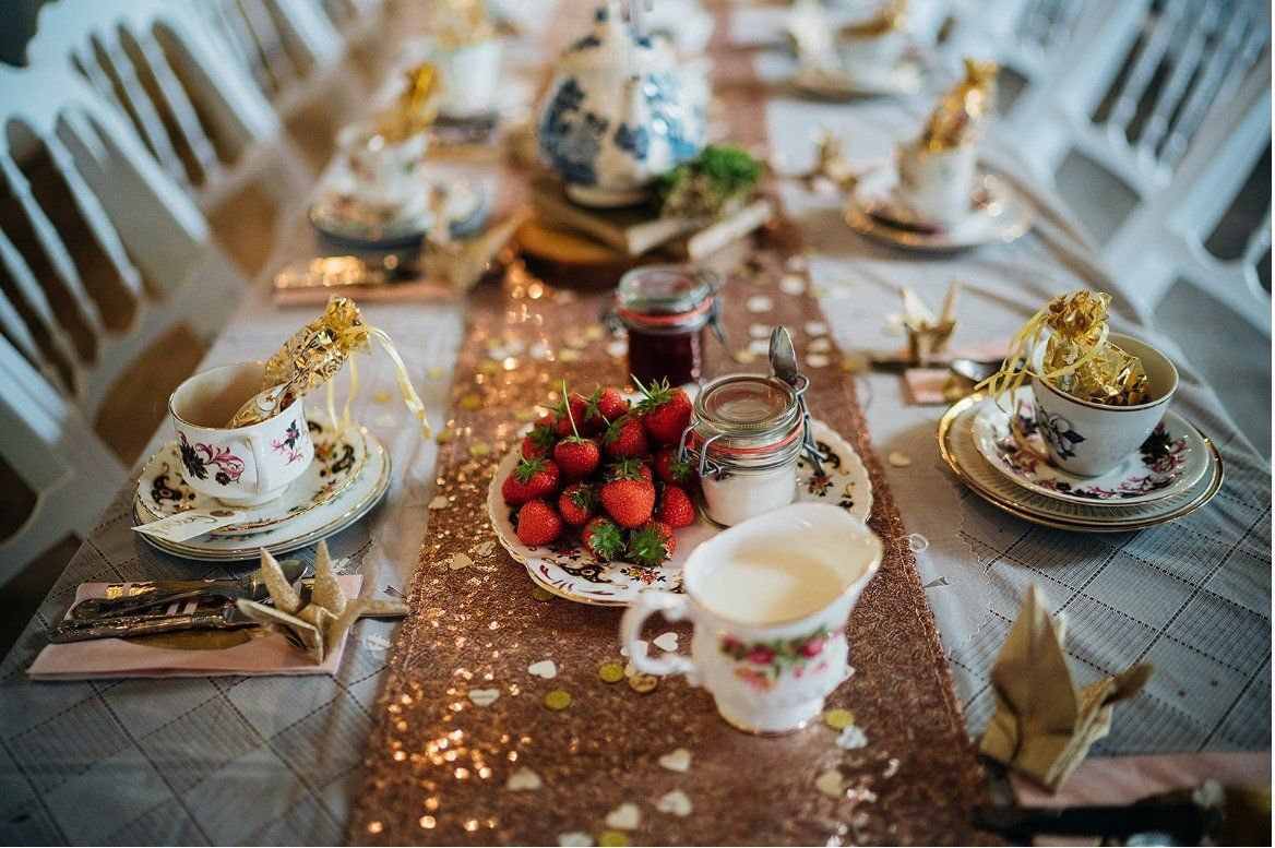 Table set for tea with strawberries, milk, and teacups on a shimmering, rose-gold runner.