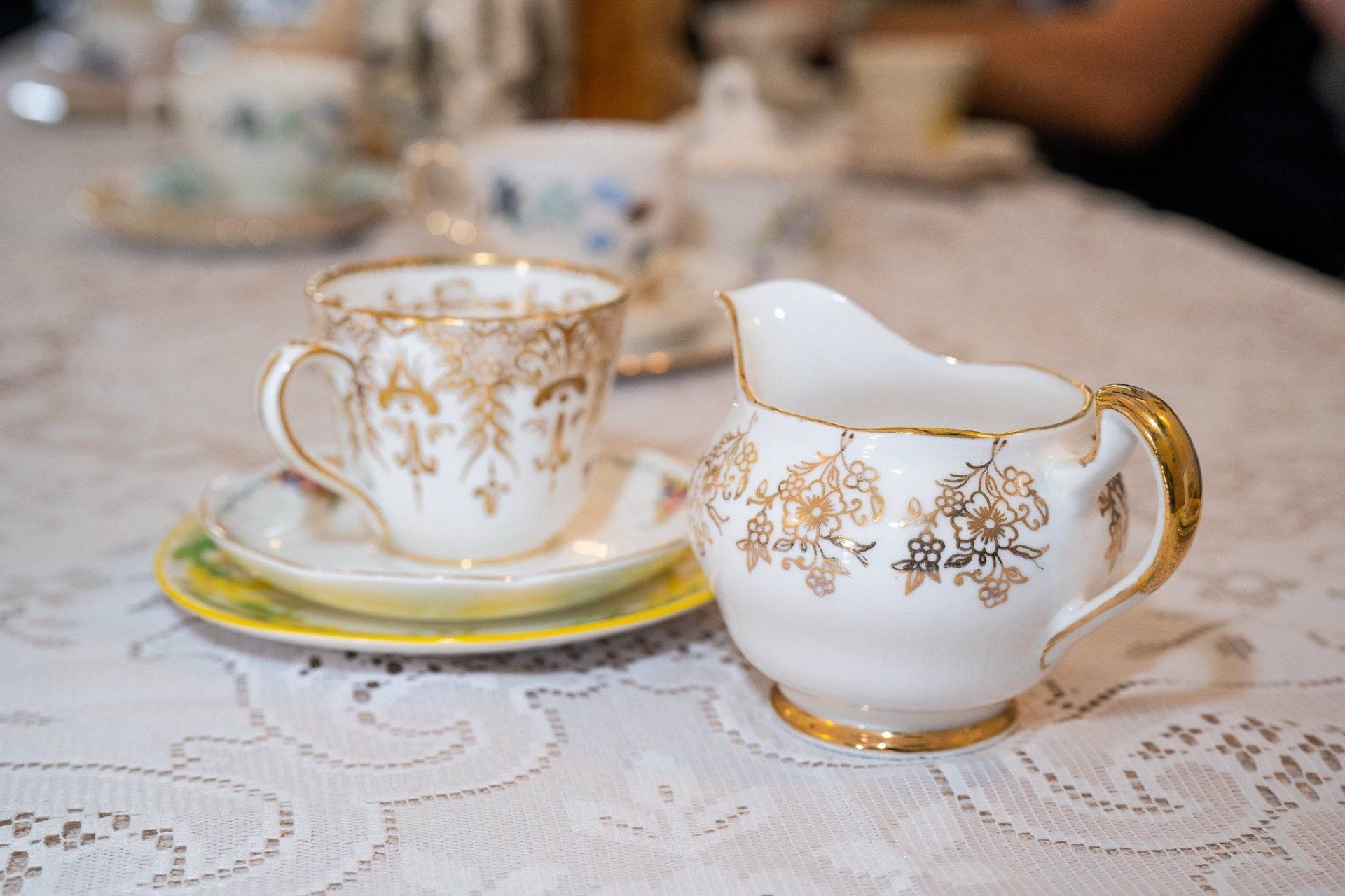 Teacup and creamer with gold embellishments on a lace-covered table.