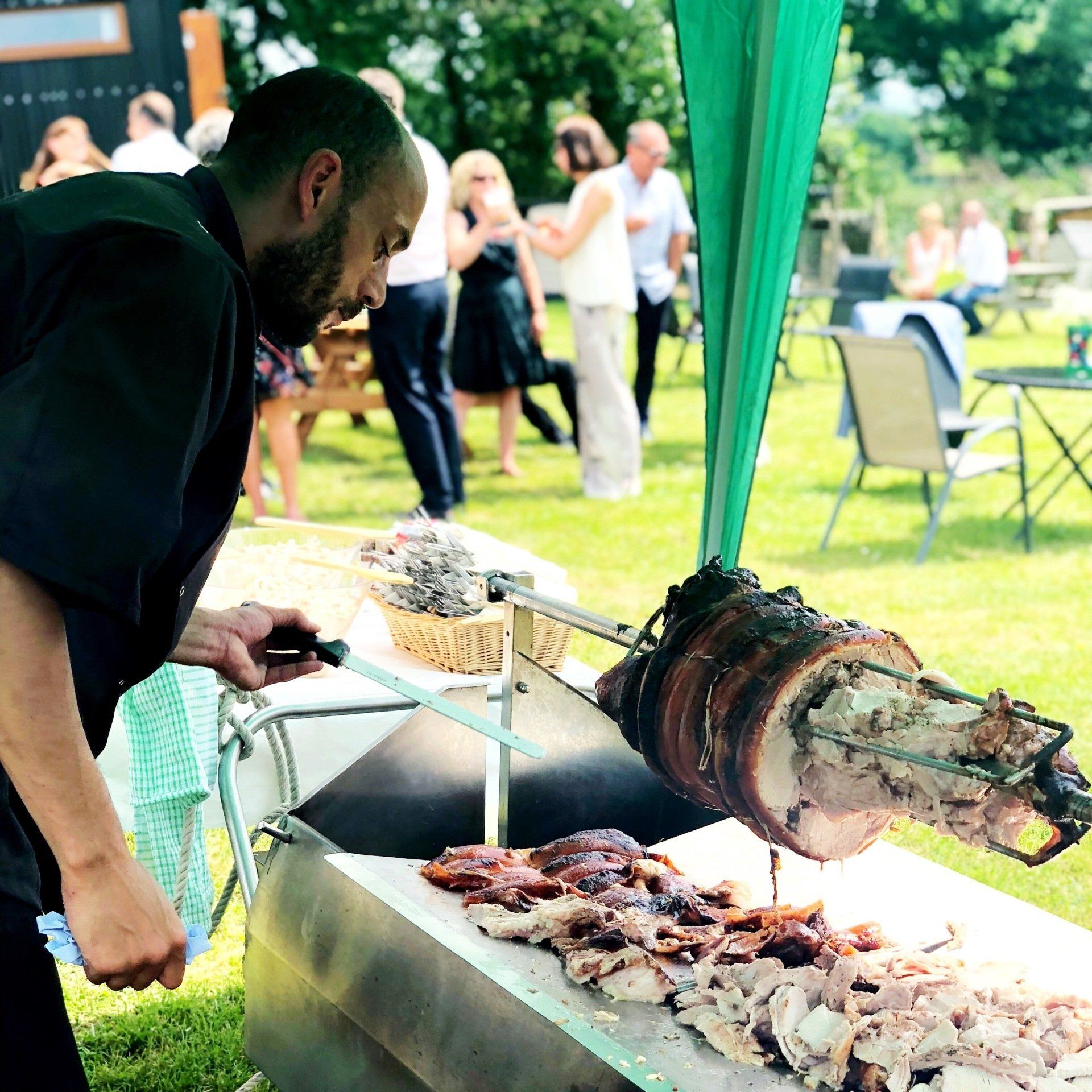 Chef carving roasted meat at an outdoor event; people and garden setting in background.
