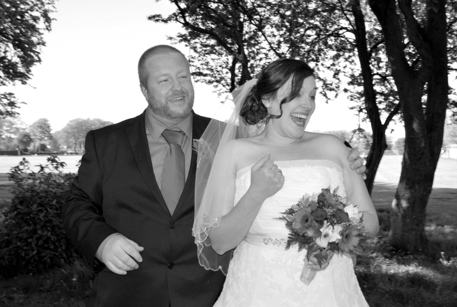 Bride and groom laughing outside, she holds bouquet, he embraces her. Black and white photo.