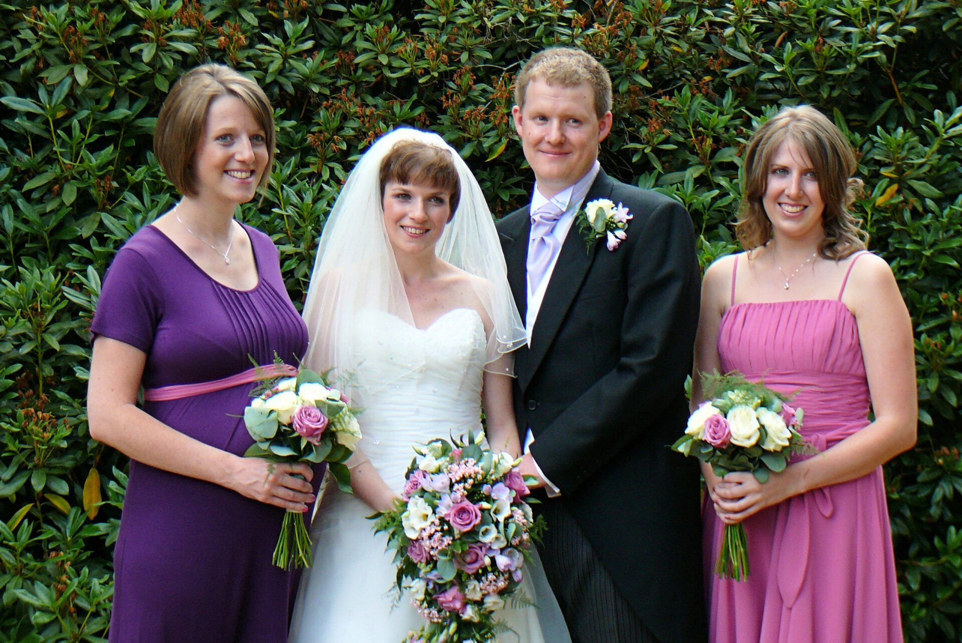 Wedding couple poses with bridesmaids in front of greenery. Bride wears white gown, groom in suit.