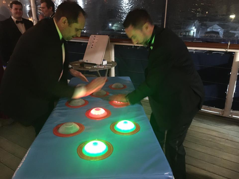 Two men in tuxedos playing a light-up game on a blue table outdoors at night.