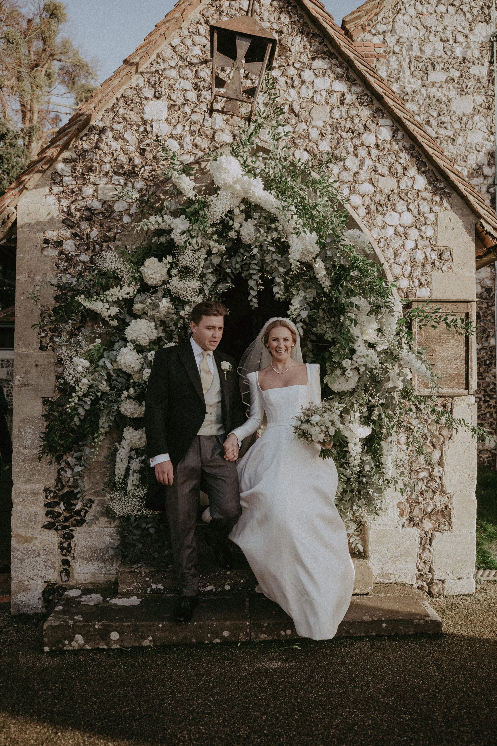 Bride and groom exiting stone church, framed by white flowers and greenery.