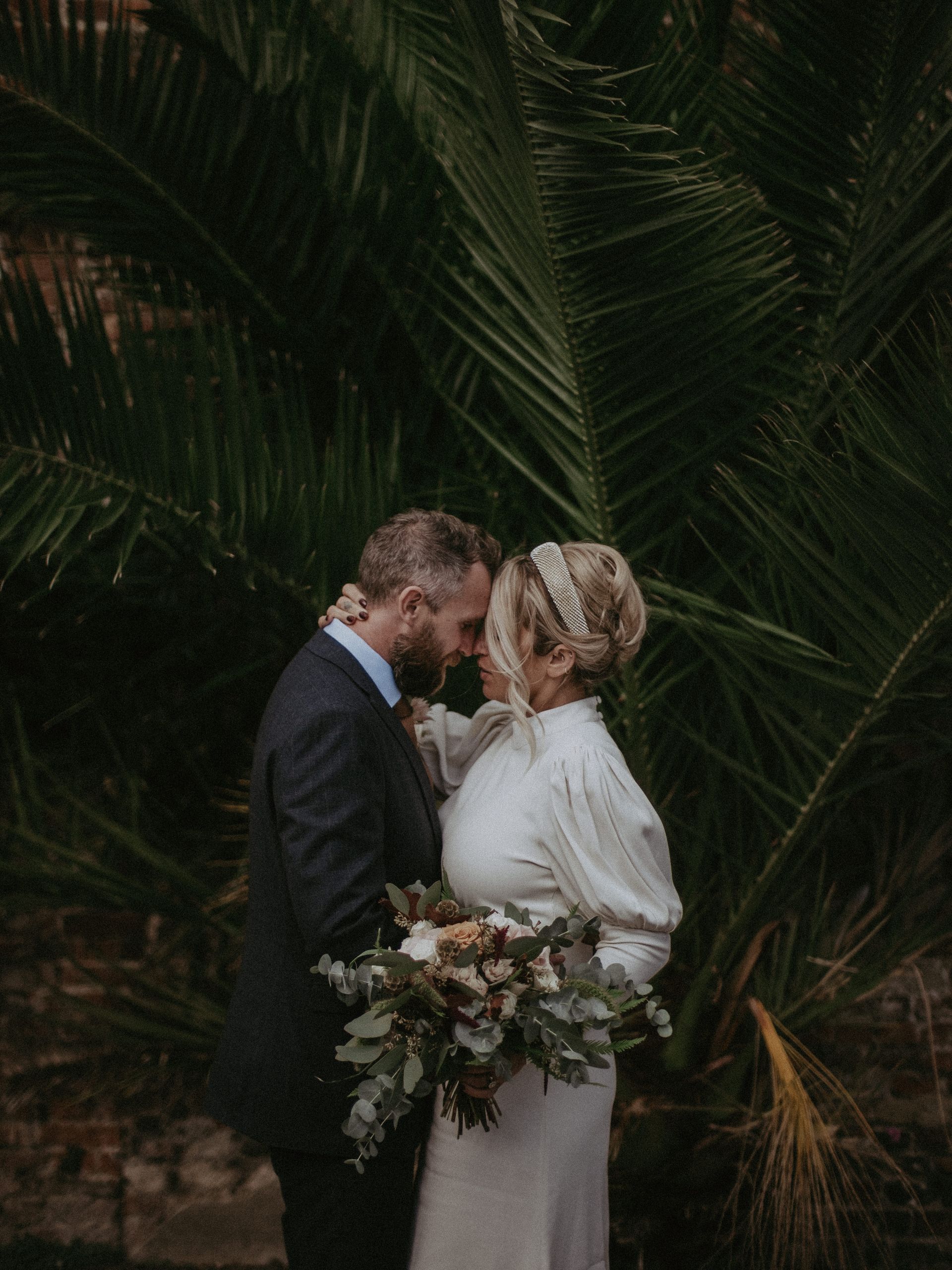 Couple embracing in front of a palm tree; the bride holds a bouquet, the groom wears a suit.