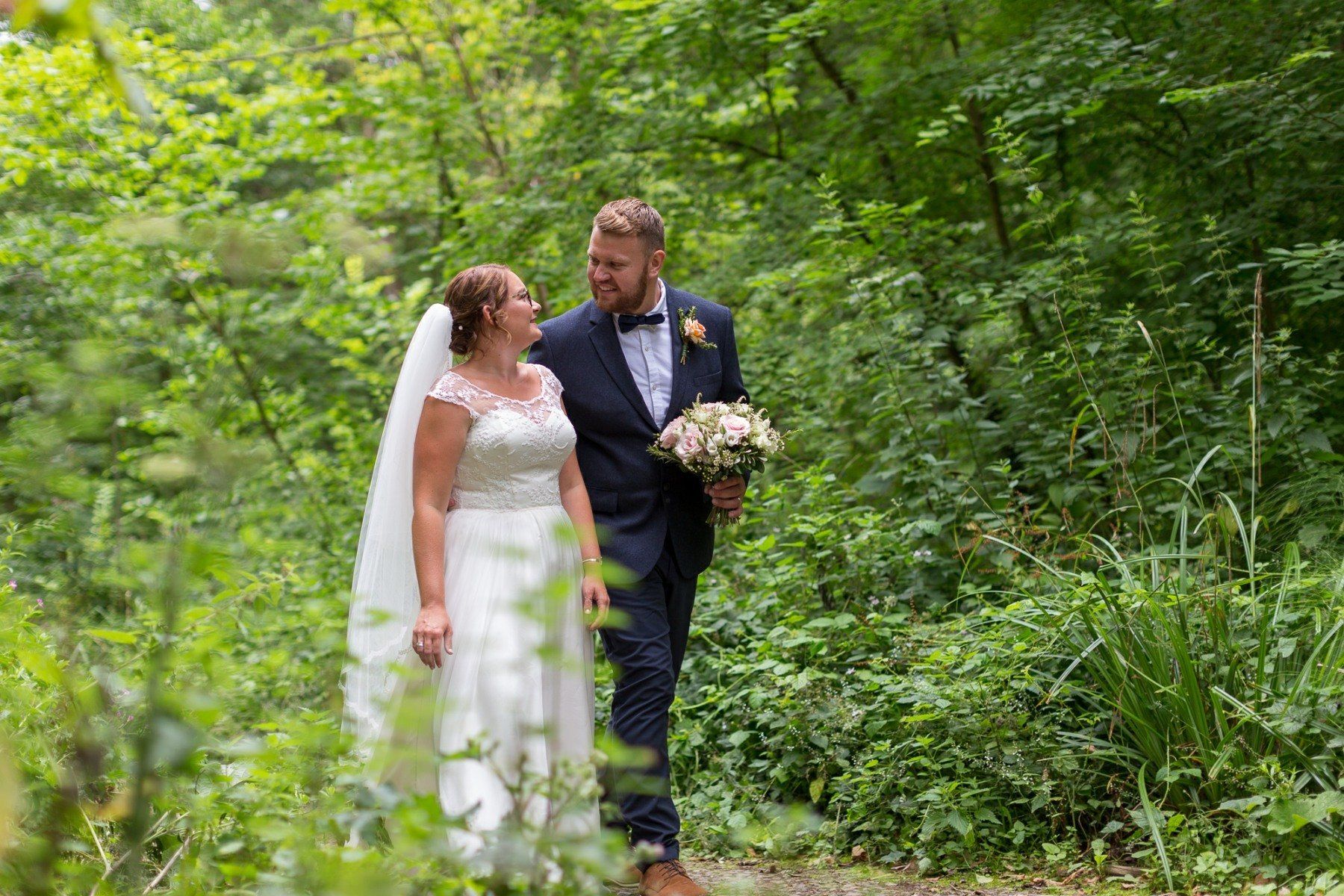 Couple walking through lush greenery; bride in white dress, groom in suit, both smiling.