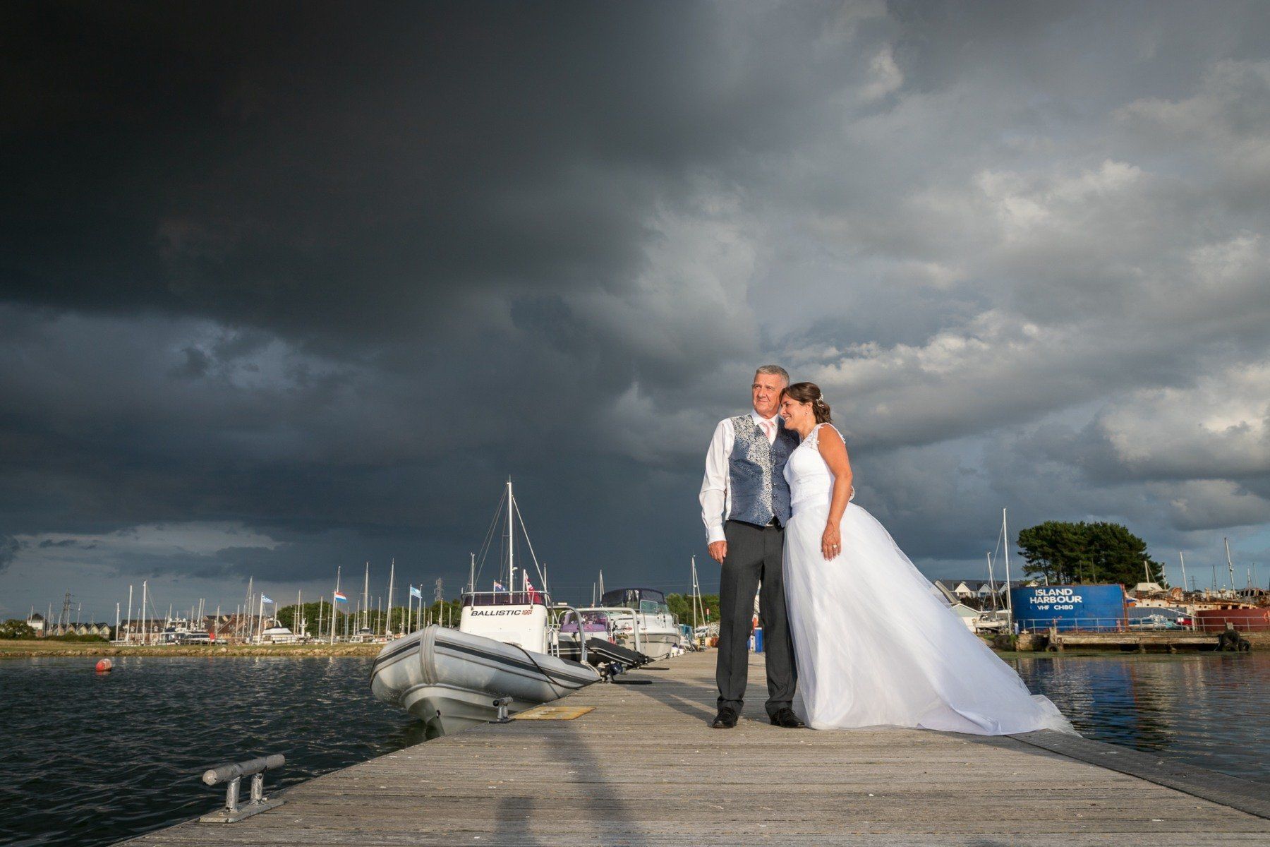 Couple on a dock with a dramatic, stormy sky, sailboats, and a boat. The bride wears a white gown.