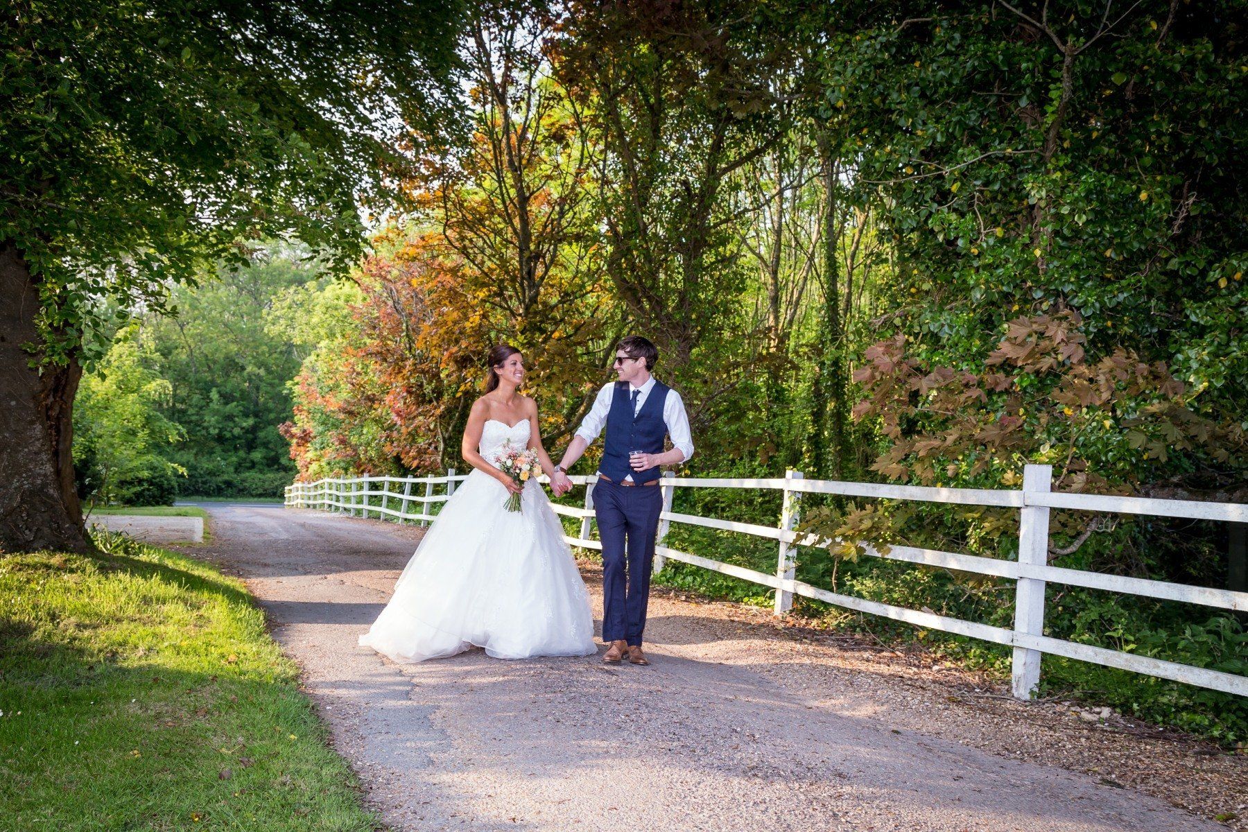 Bride and groom walk hand-in-hand on a gravel path, past white fence and trees, wedding day.