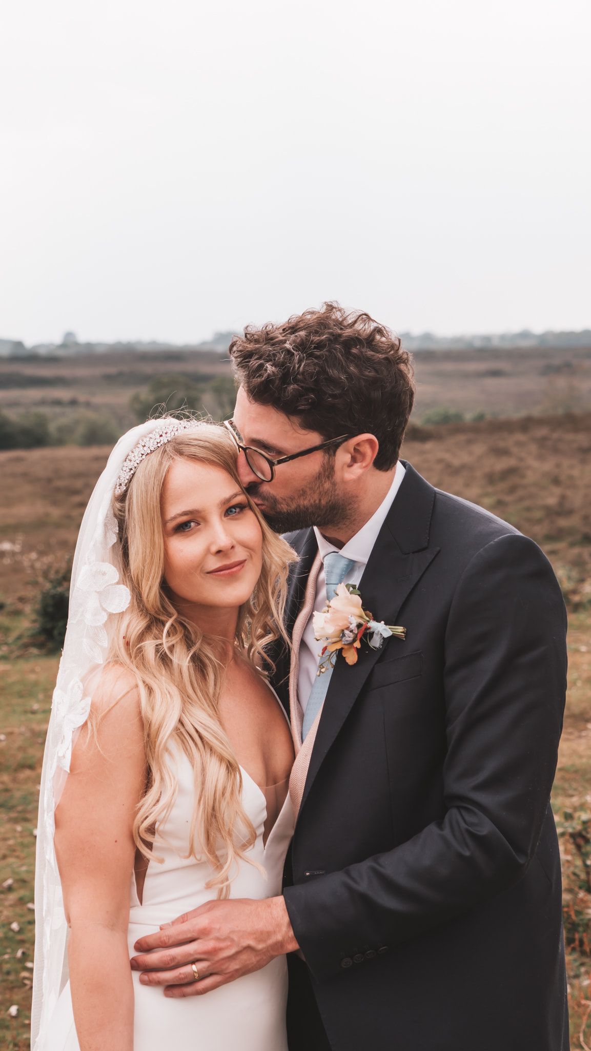 Bride in a white dress and veil with groom in a suit kissing her forehead in an outdoor setting.