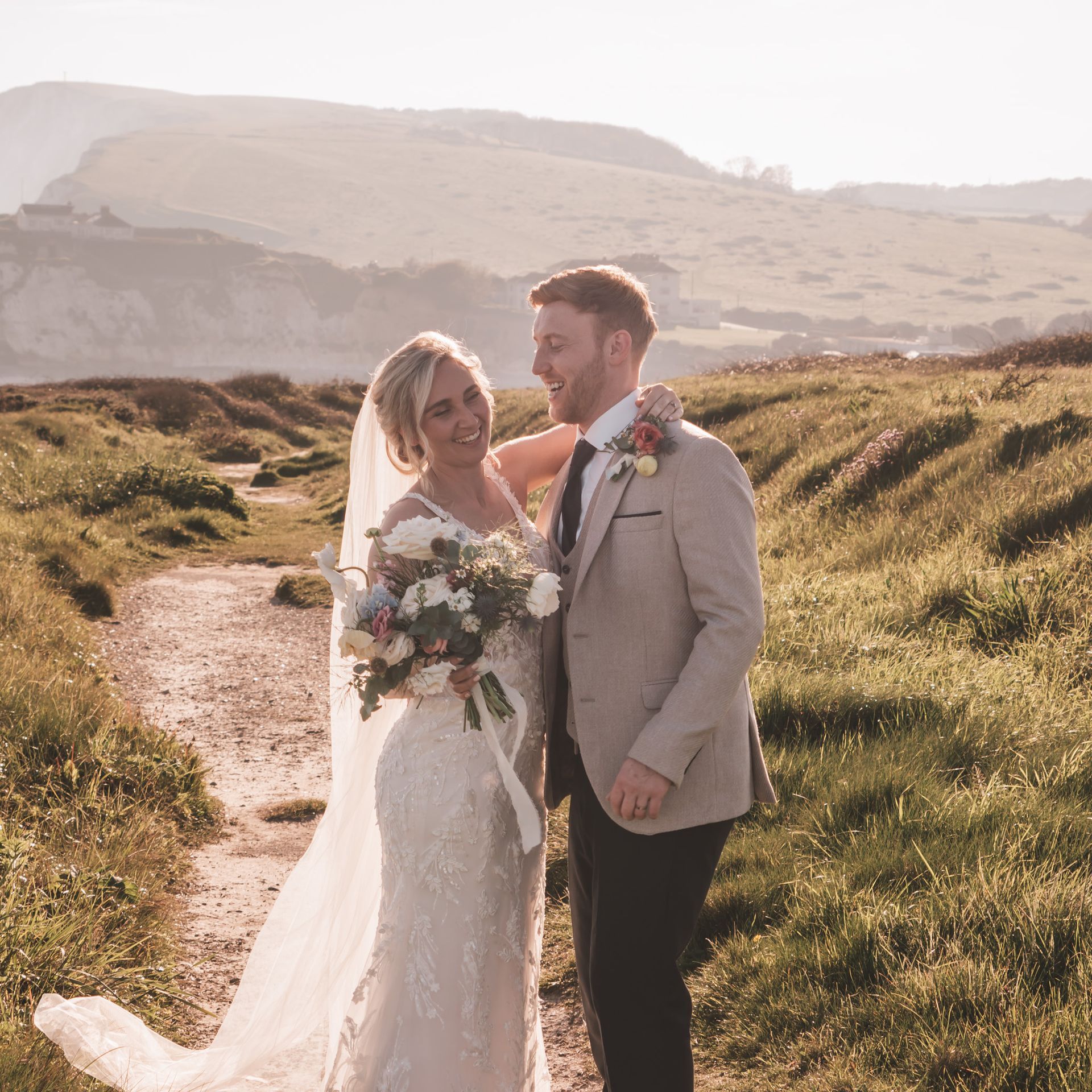 Bride and groom smiling, posing for photo on path with hillside background.