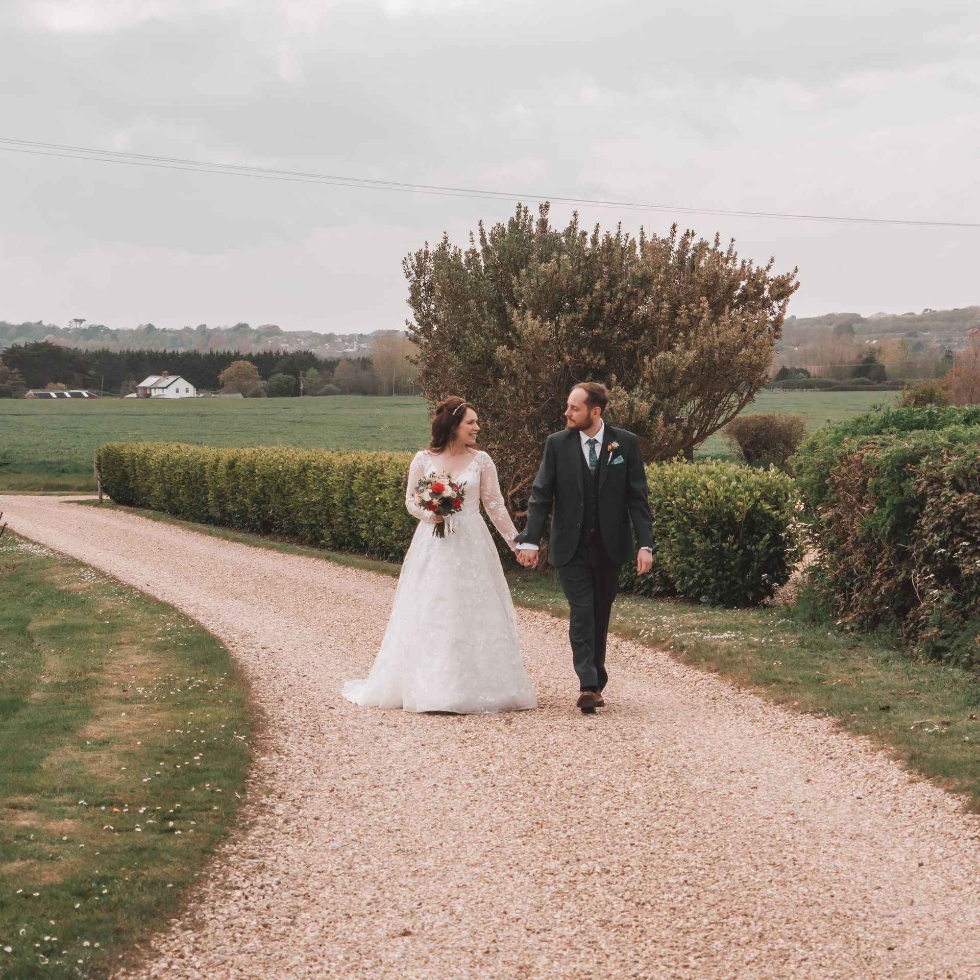 Bride and groom walking hand-in-hand on a gravel path, surrounded by greenery. Overcast day.