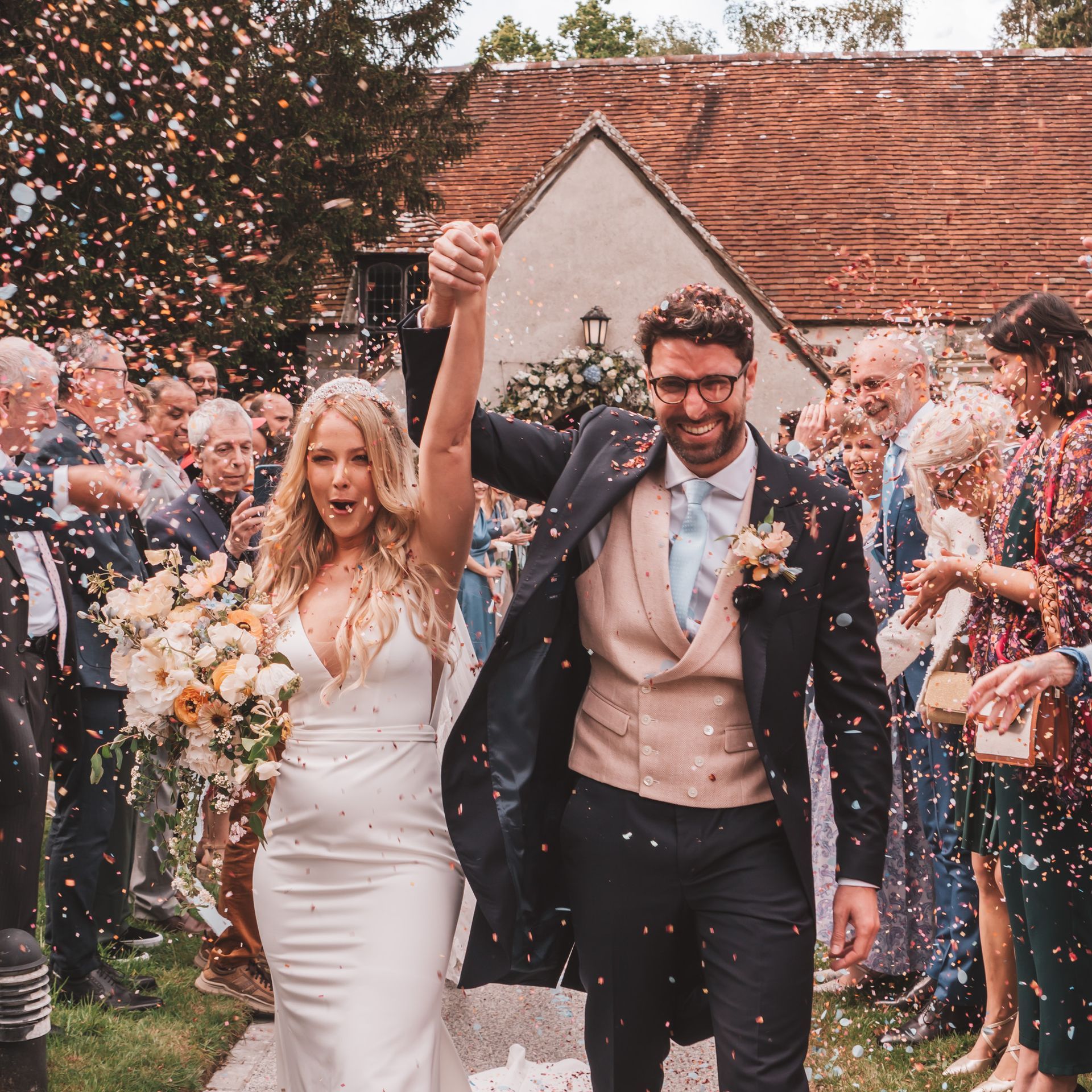 Newlyweds celebrate, walking through confetti with guests; couple smiles, he in suit and she in white dress.