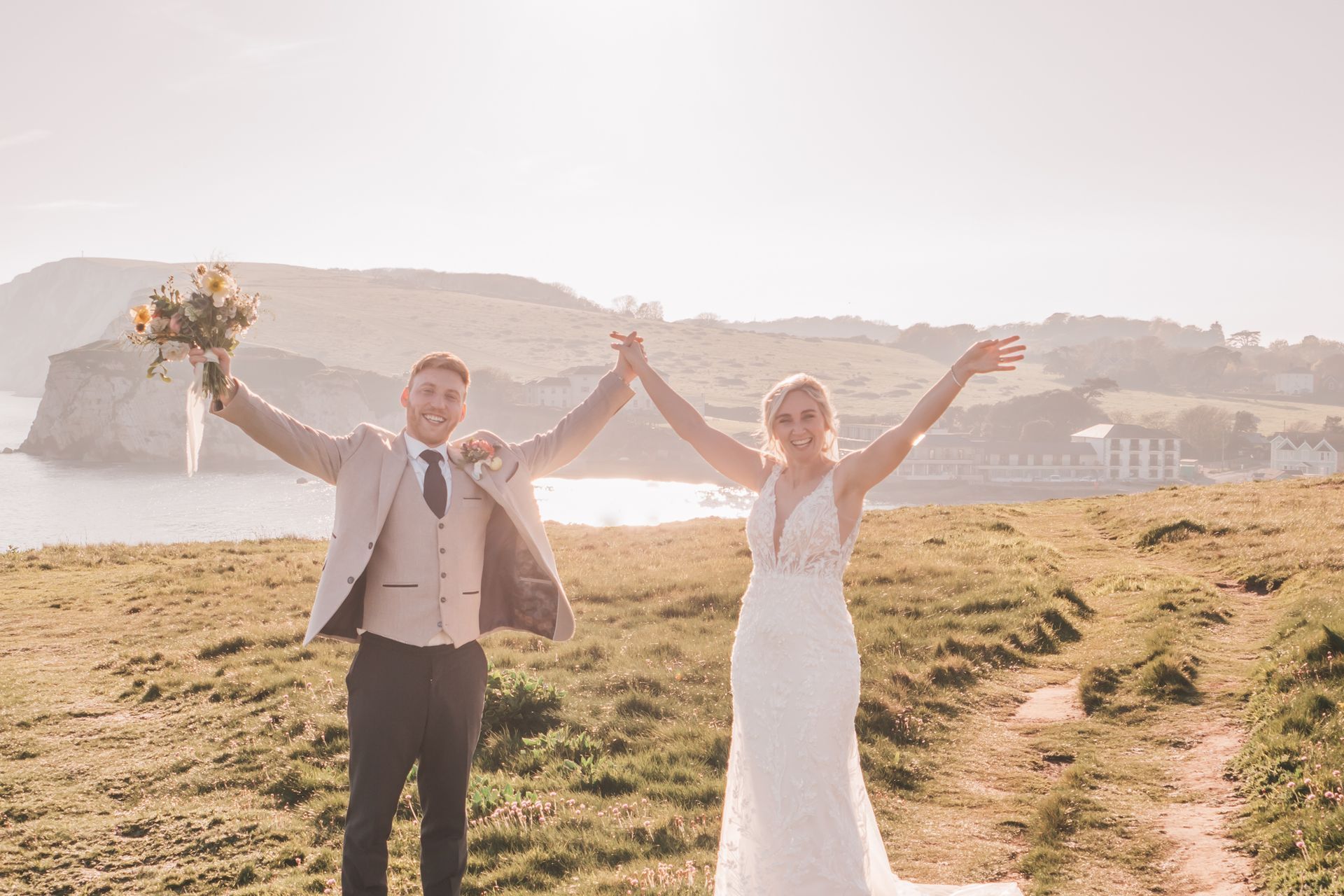 Couple holding hands, arms raised in celebration on a cliff overlooking the sea; sunny, joyful.