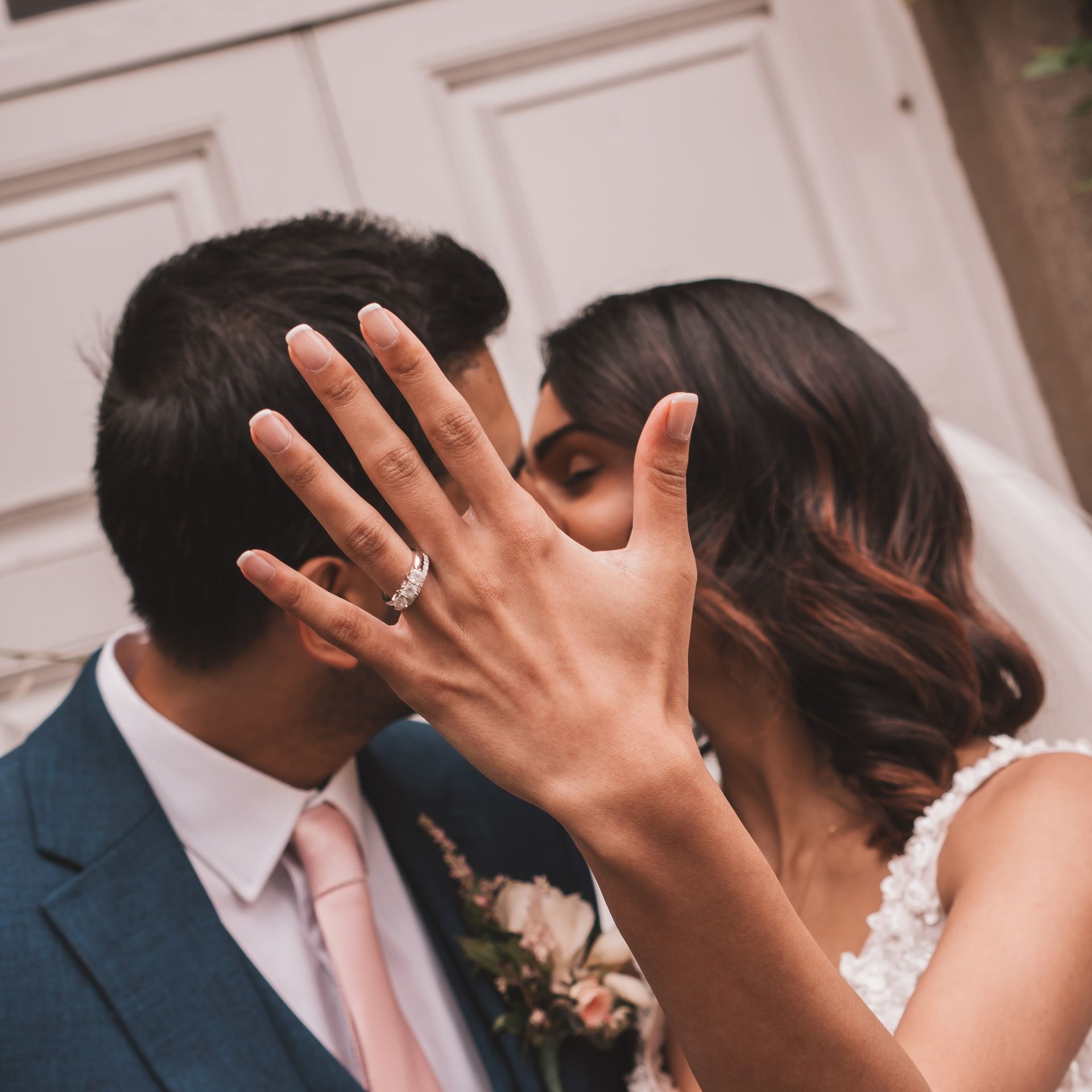 Newlyweds kissing, bride showing off diamond ring, against white doors, pink tie, blue suit.