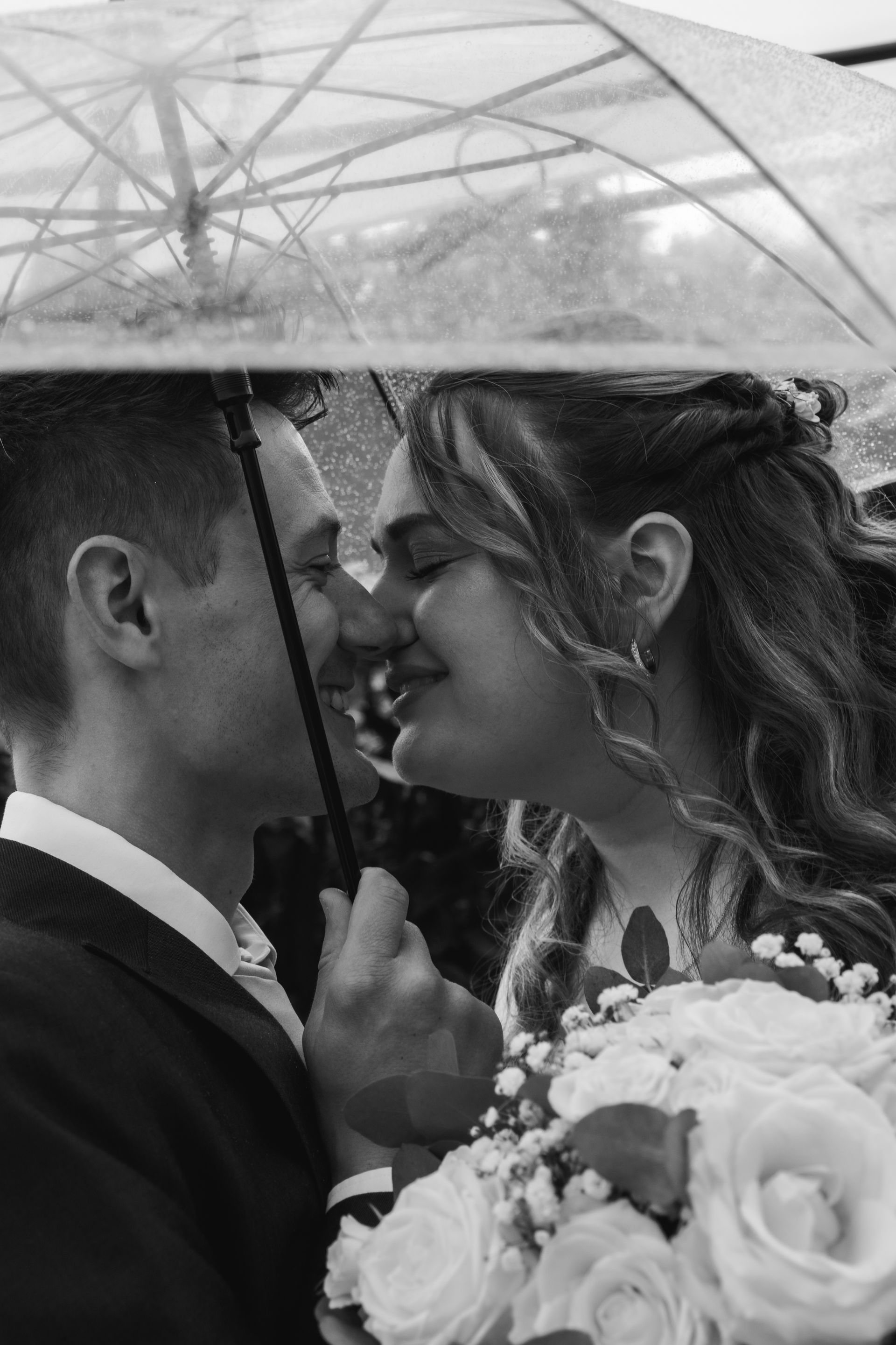 Couple sharing a kiss under a clear umbrella, wedding bouquet visible. Black and white.