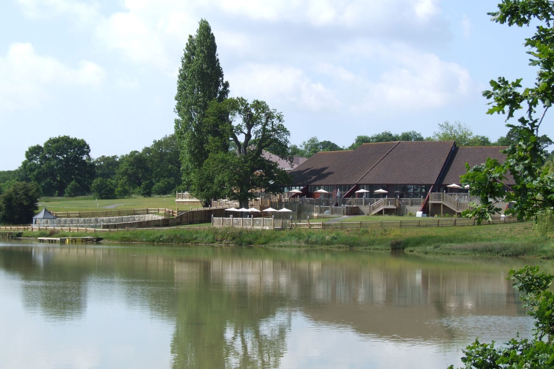 Lake with a building, tall trees, and wooden structure on the edge. Sunny day with reflections.