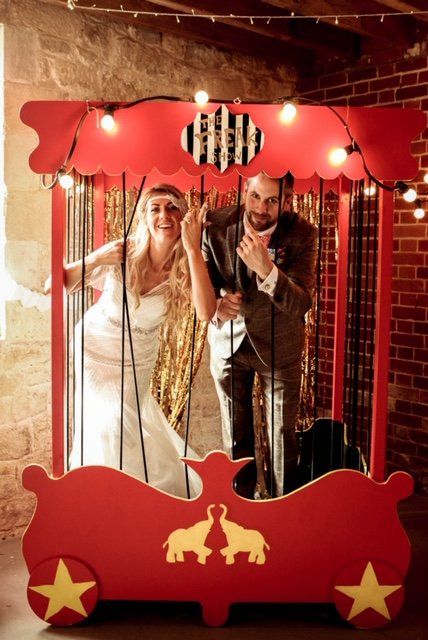Bride and groom in circus-themed photo booth. Red frame with lights, gold fringe backdrop. Smiles and poses.