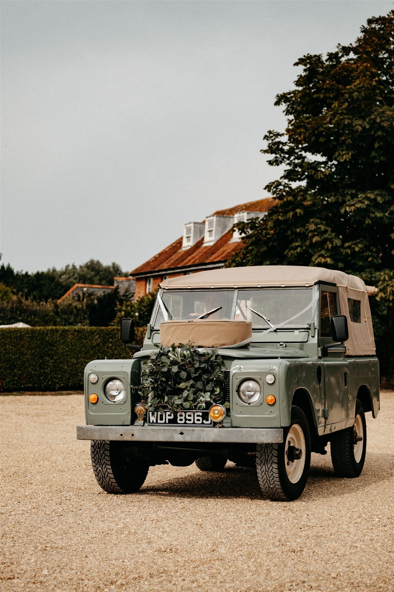 Green Land Rover parked on gravel in front of a building with a green hedge and trees.