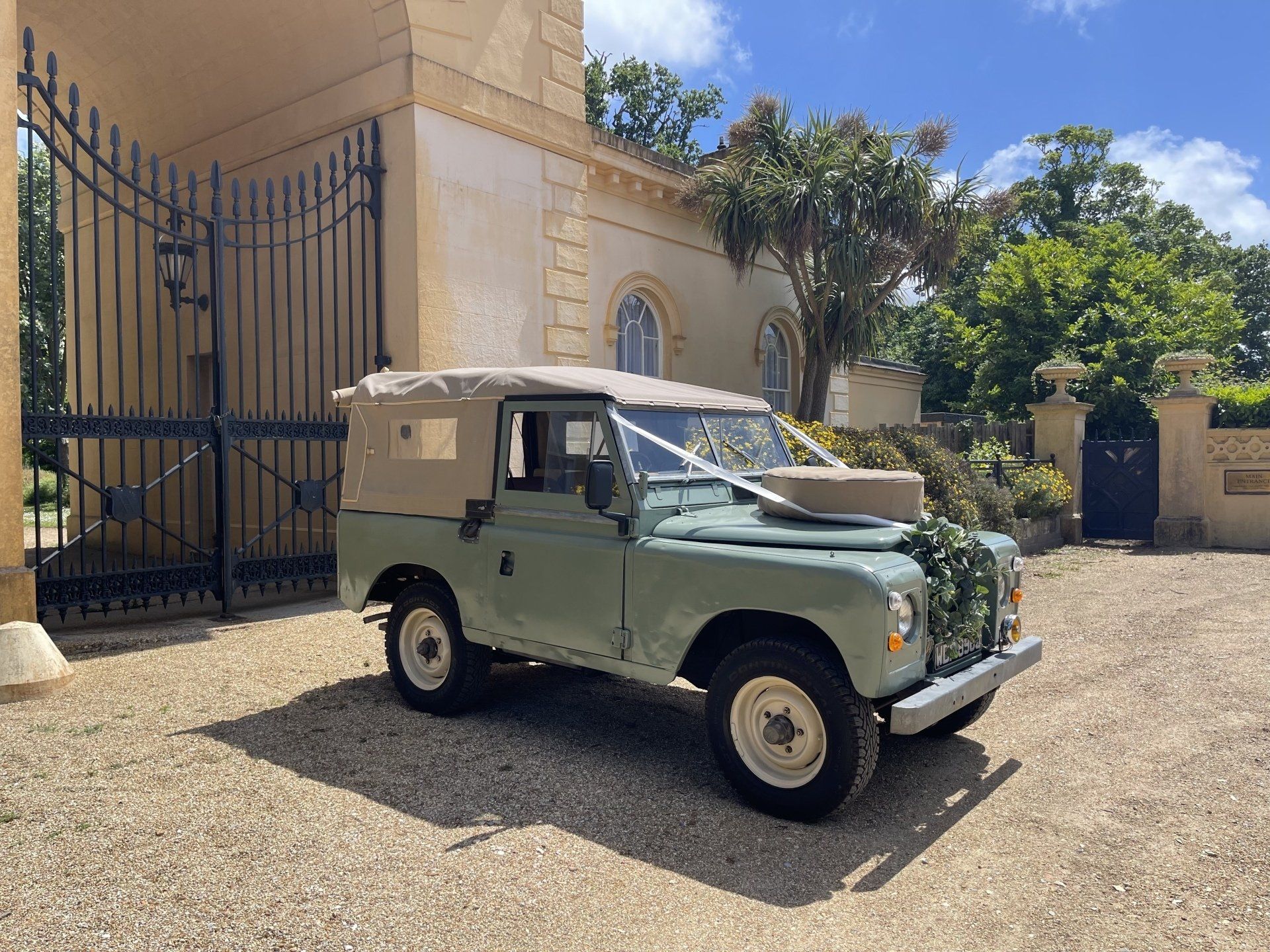 Light green vintage Land Rover parked in front of a stone building with black gates; decorated for a wedding.