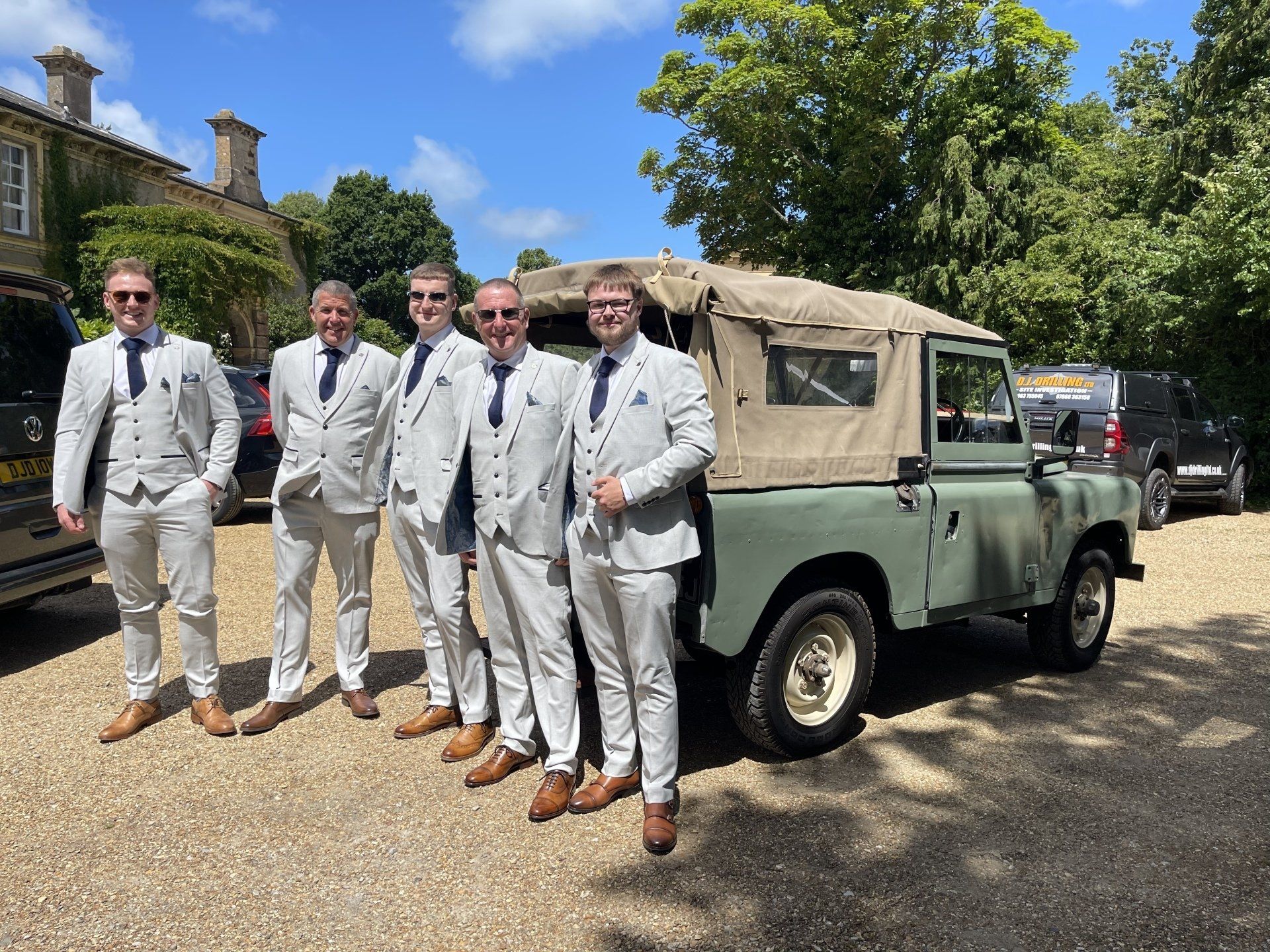 Five men in light gray suits pose next to an olive green vintage Land Rover on a sunny day.