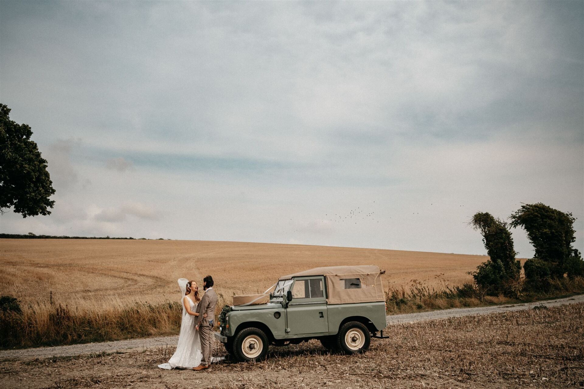Couple kissing by a vintage green Land Rover in a field under a cloudy sky.