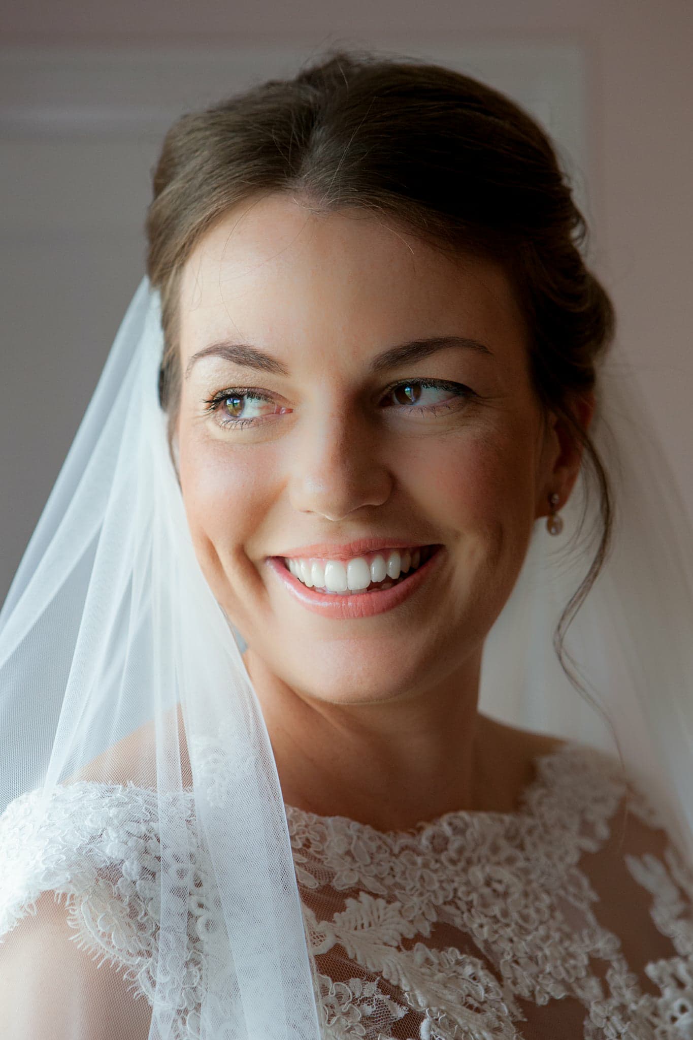 Bride in white lace dress and veil, smiling with updo hairstyle; soft pink background.