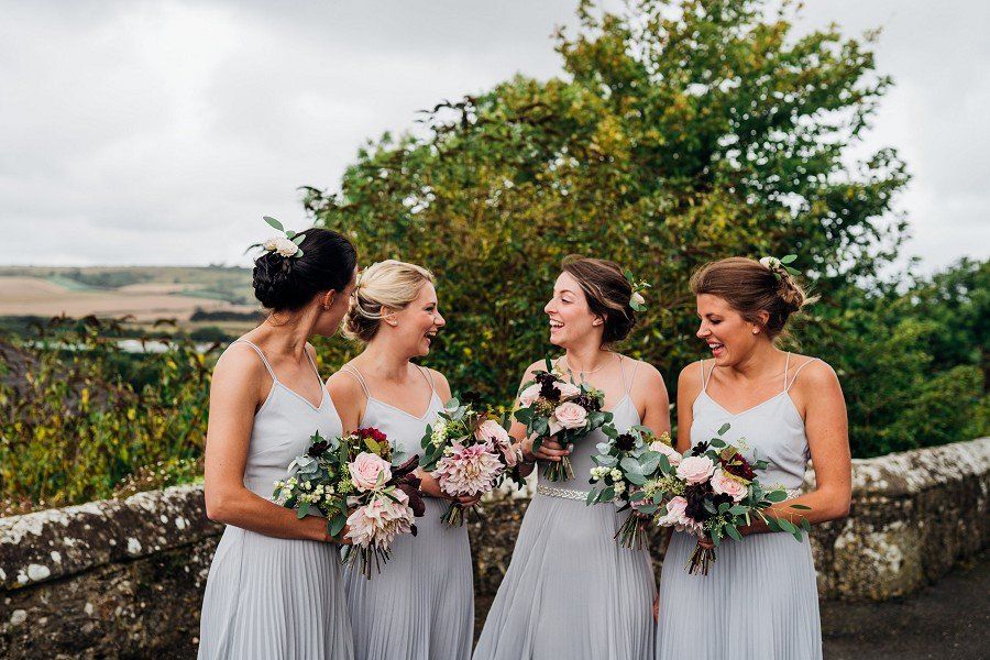 Bridesmaids in light blue dresses holding bouquets, laughing outdoors near a stone wall.