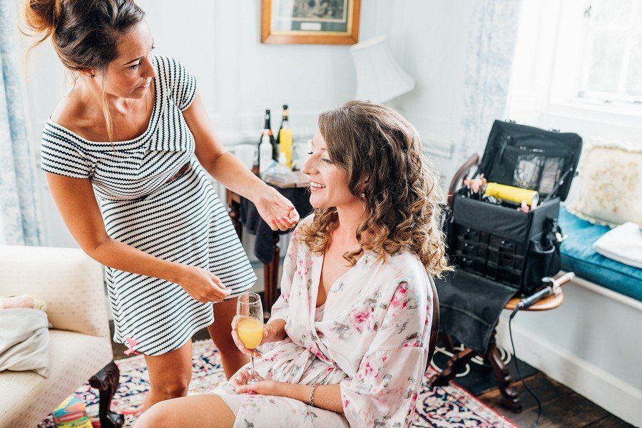 Woman in floral robe has hair styled; another woman adjusts her hair, with makeup case nearby.
