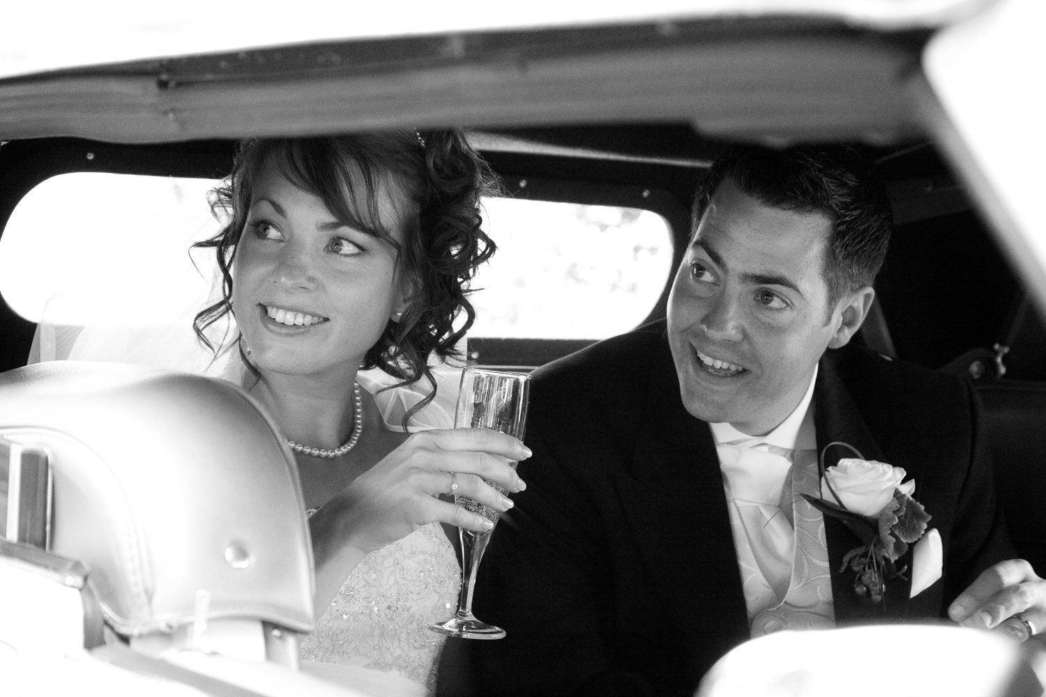 Bride and groom smiling inside a wedding car, the bride holding a champagne flute.