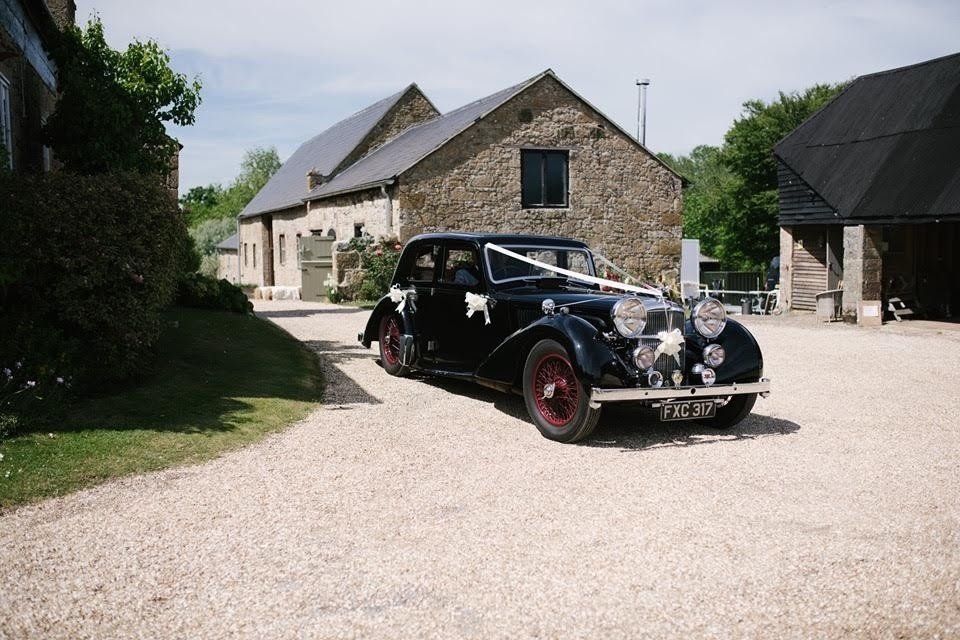 Black vintage car with ribbons parked in front of a stone building with a gravel driveway.