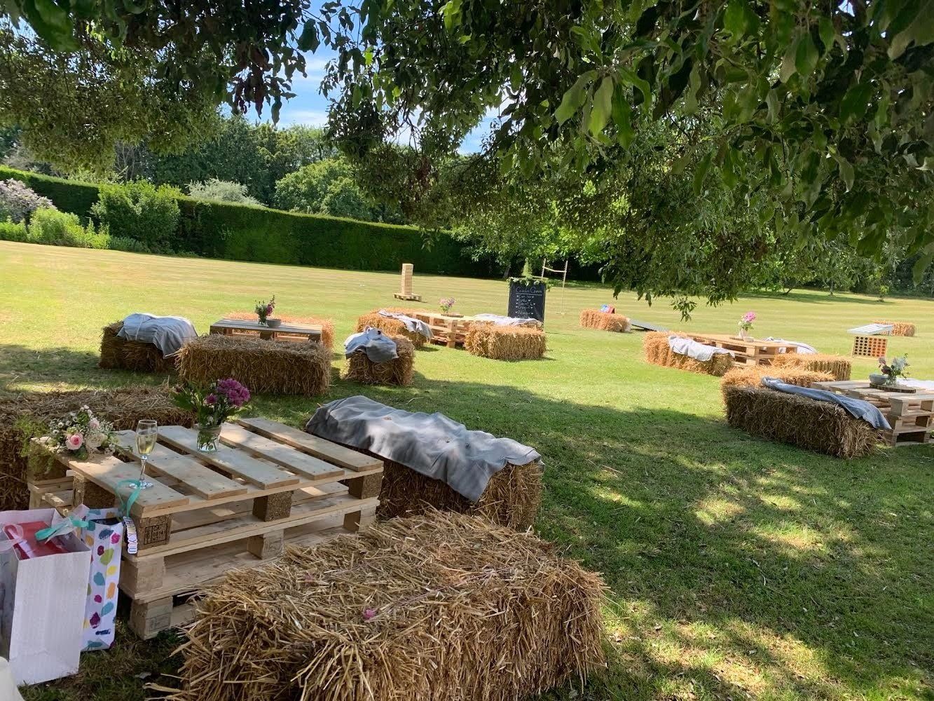 Outdoor seating area with hay bales and pallet tables on grass under trees.