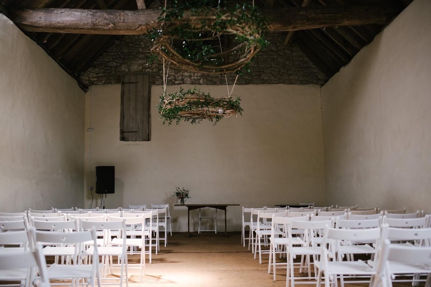 Wedding ceremony in a rustic barn, white chairs arranged in rows. Floral wreaths hang from the ceiling.