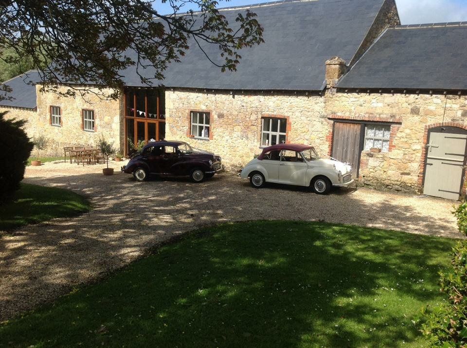 Two vintage cars parked in front of a stone building with a gravel driveway and green lawn.