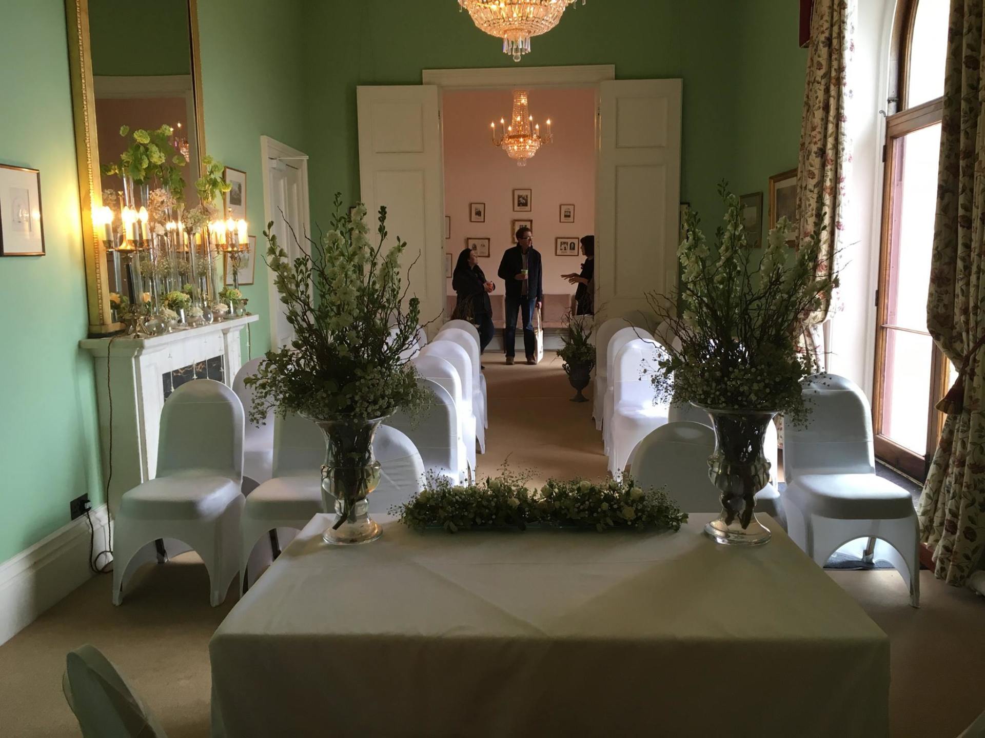 Wedding ceremony setup: table, chairs with white covers, floral arrangements, and guests in a light green room.
