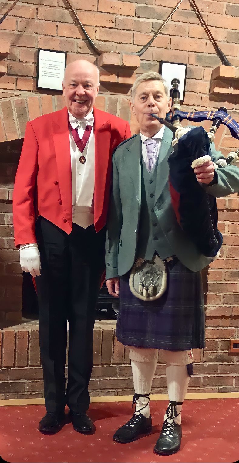Two men: One in formal red coat, one in Scottish attire, including a kilt and bagpipes, pose near a brick wall.