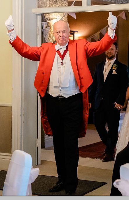Man in red coat with white gloves, arms raised, standing in doorway at event.