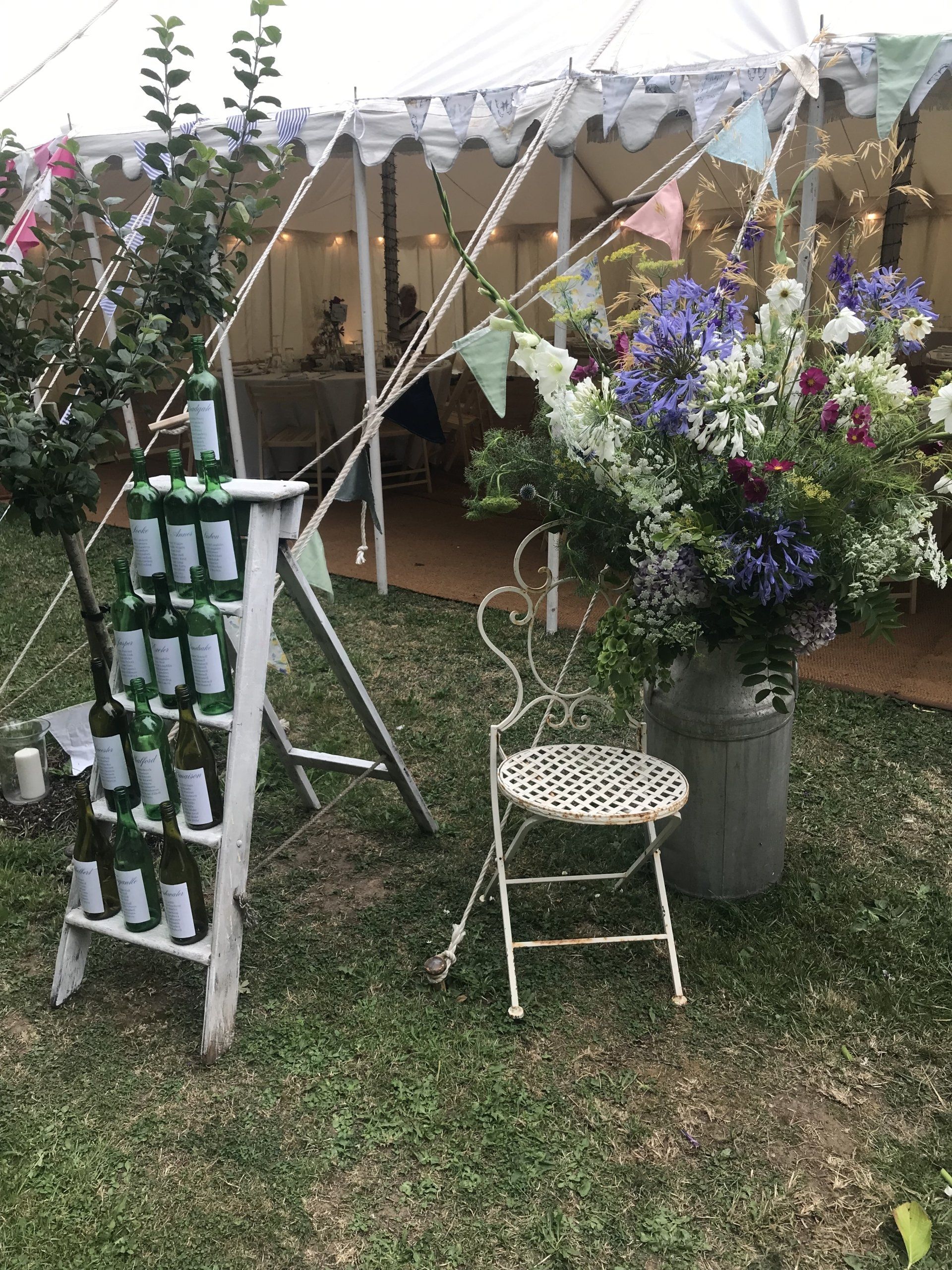 Wedding decor with a tent backdrop, featuring a floral display, a bottle display, and a decorative chair on a grassy lawn.