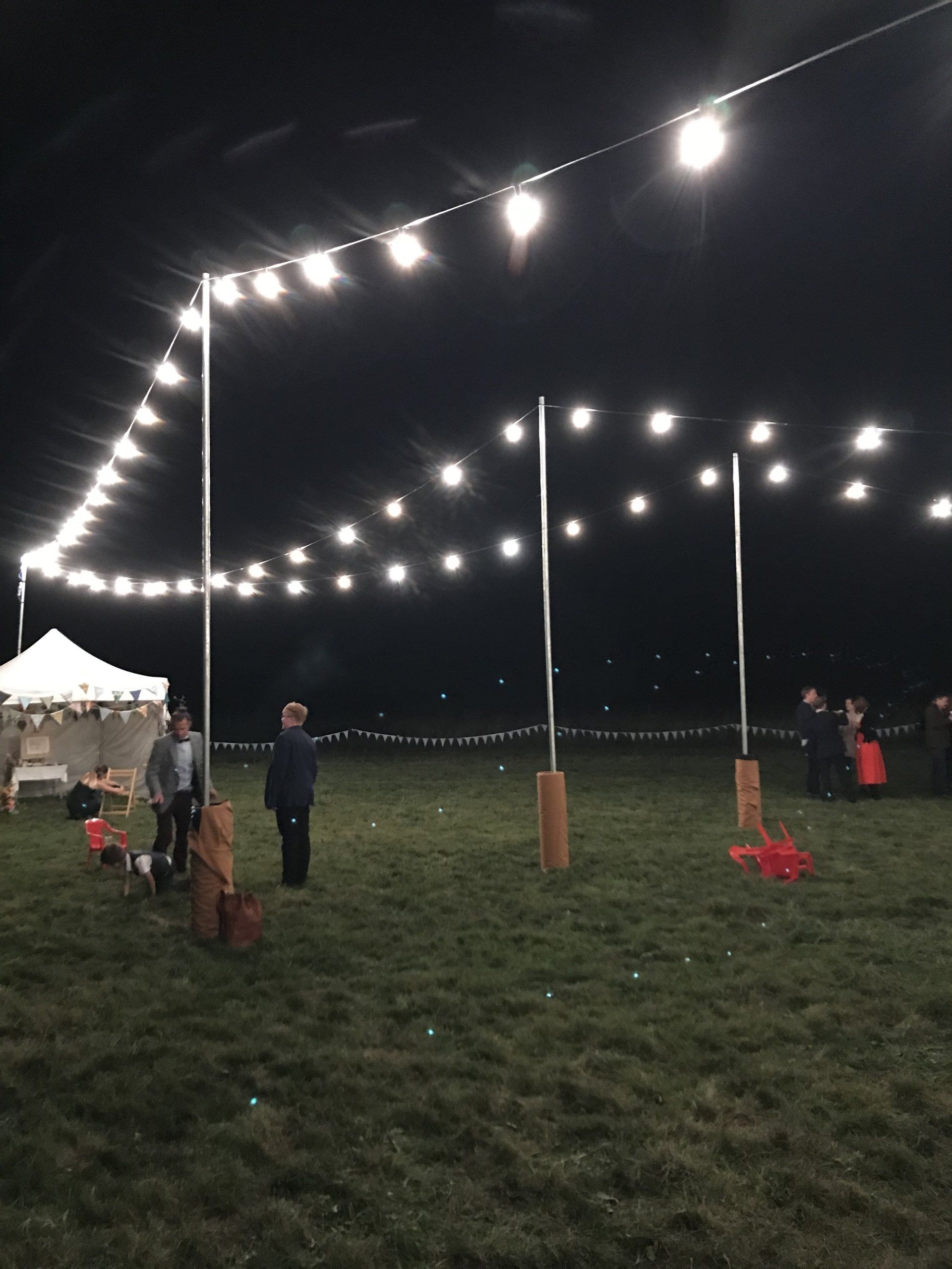String lights illuminate a grassy field at night. Three poles support the lights, and people stand nearby. A tent is visible.