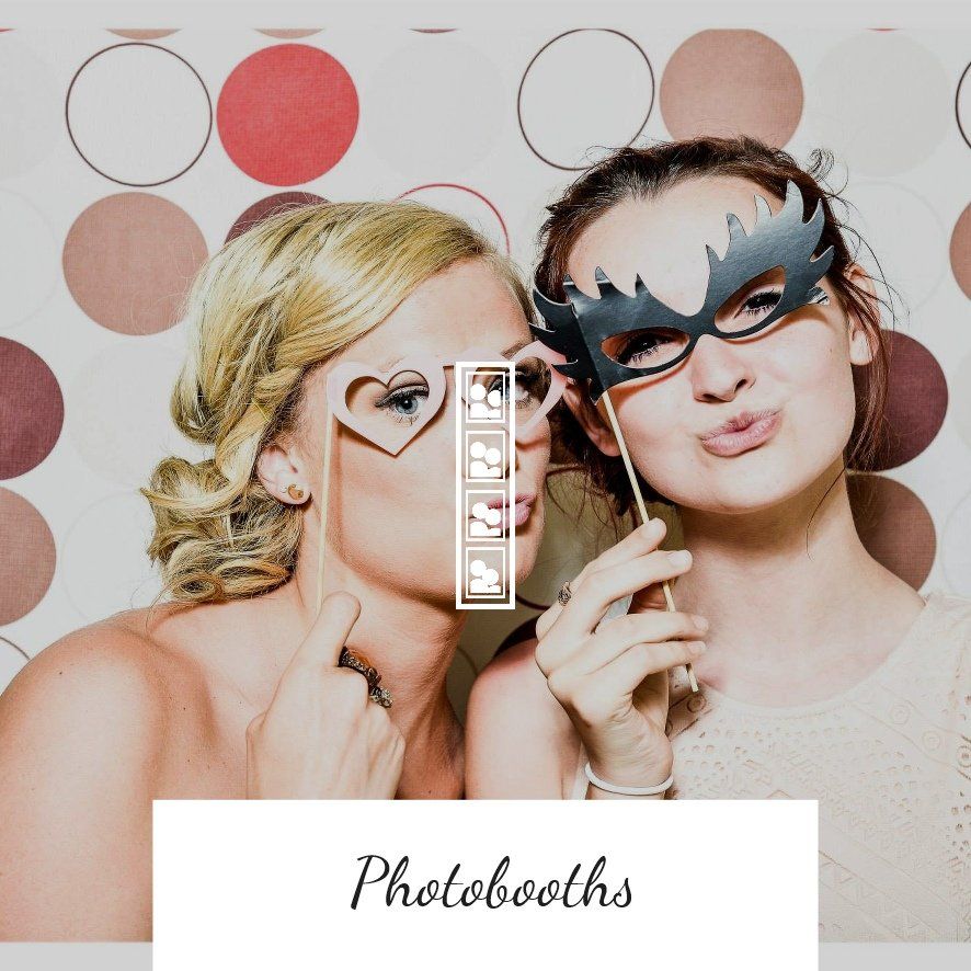 Two women with masks posing for a photo booth. One holds a heart-shaped mask, the other a bird-like mask. Polka dot backdrop.