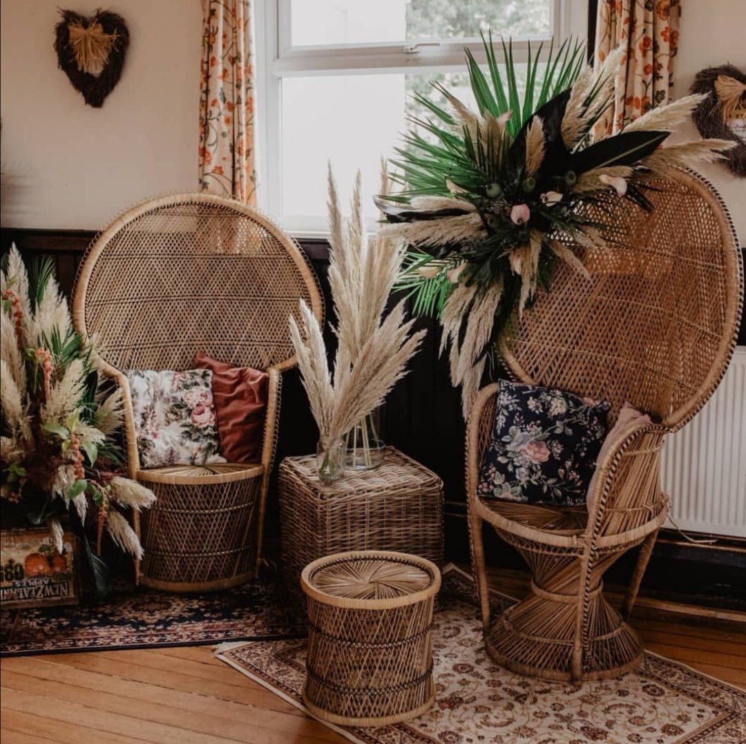 Two peacock chairs, wicker table, pampas grass, floral pillows, ornate rug, and window in a room.